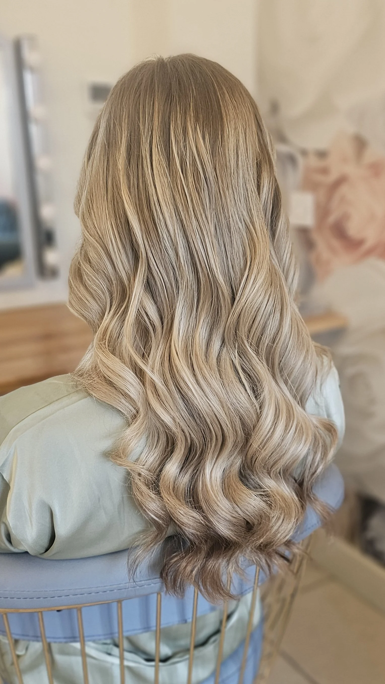 Back view of a woman with long, wavy, blonde hair sitting on a blue chair in a room with a blurred background.