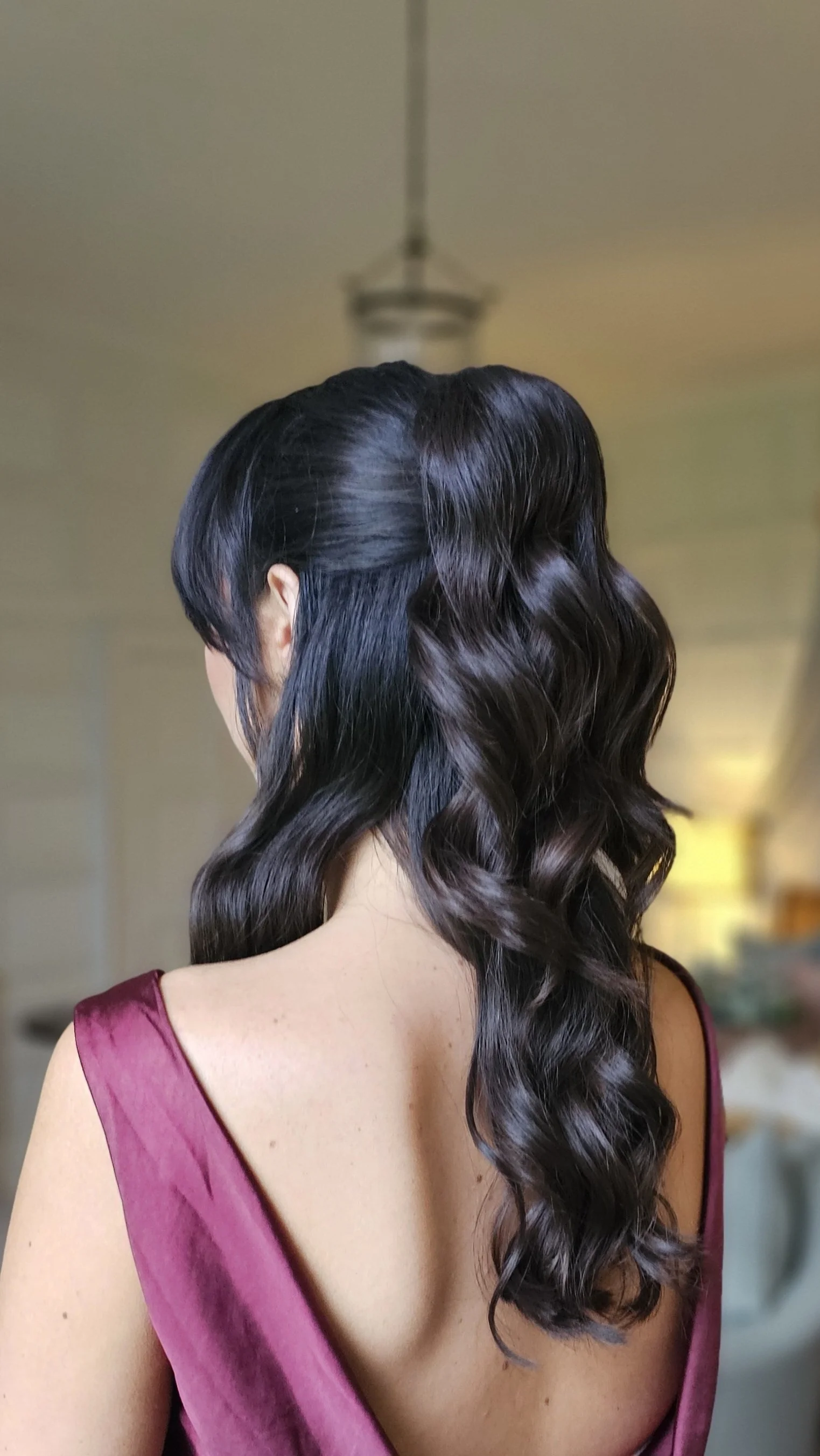 Woman with long, wavy dark hair in a maroon satin dress, viewed from behind in a softly lit room.