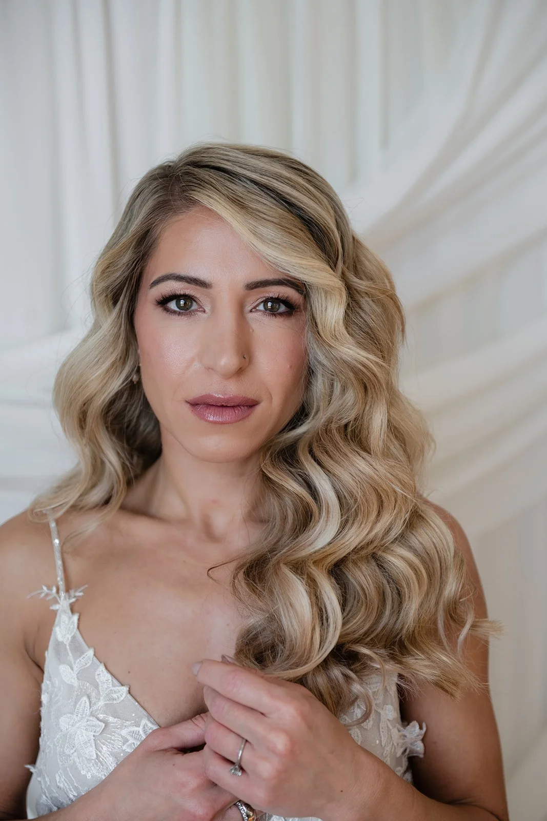 A woman with long, blonde, wavy hair and light makeup, wearing a white lace dress, is looking directly at the camera against a light-colored background.