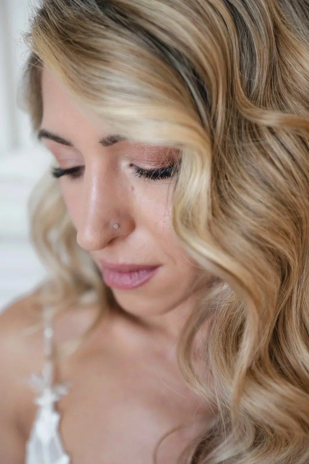 Close-up of a smiling woman with long, wavy blonde hair, a nose piercing, and wearing makeup in soft pink tones.