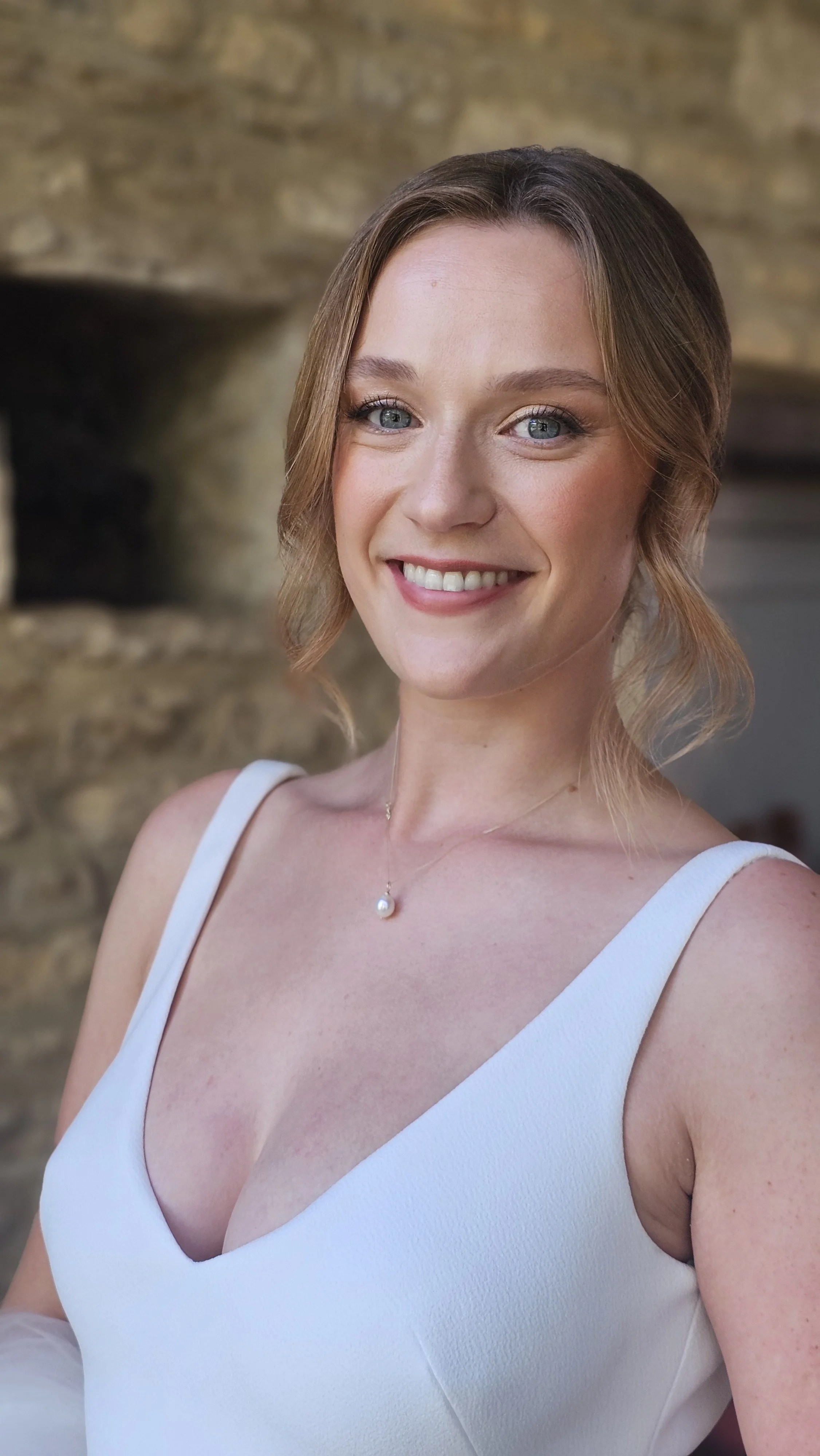 A smiling woman with light brown hair styled in loose waves, wearing a white sleeveless dress with a V-neckline and a pearl necklace. She is standing in front of a stone background.