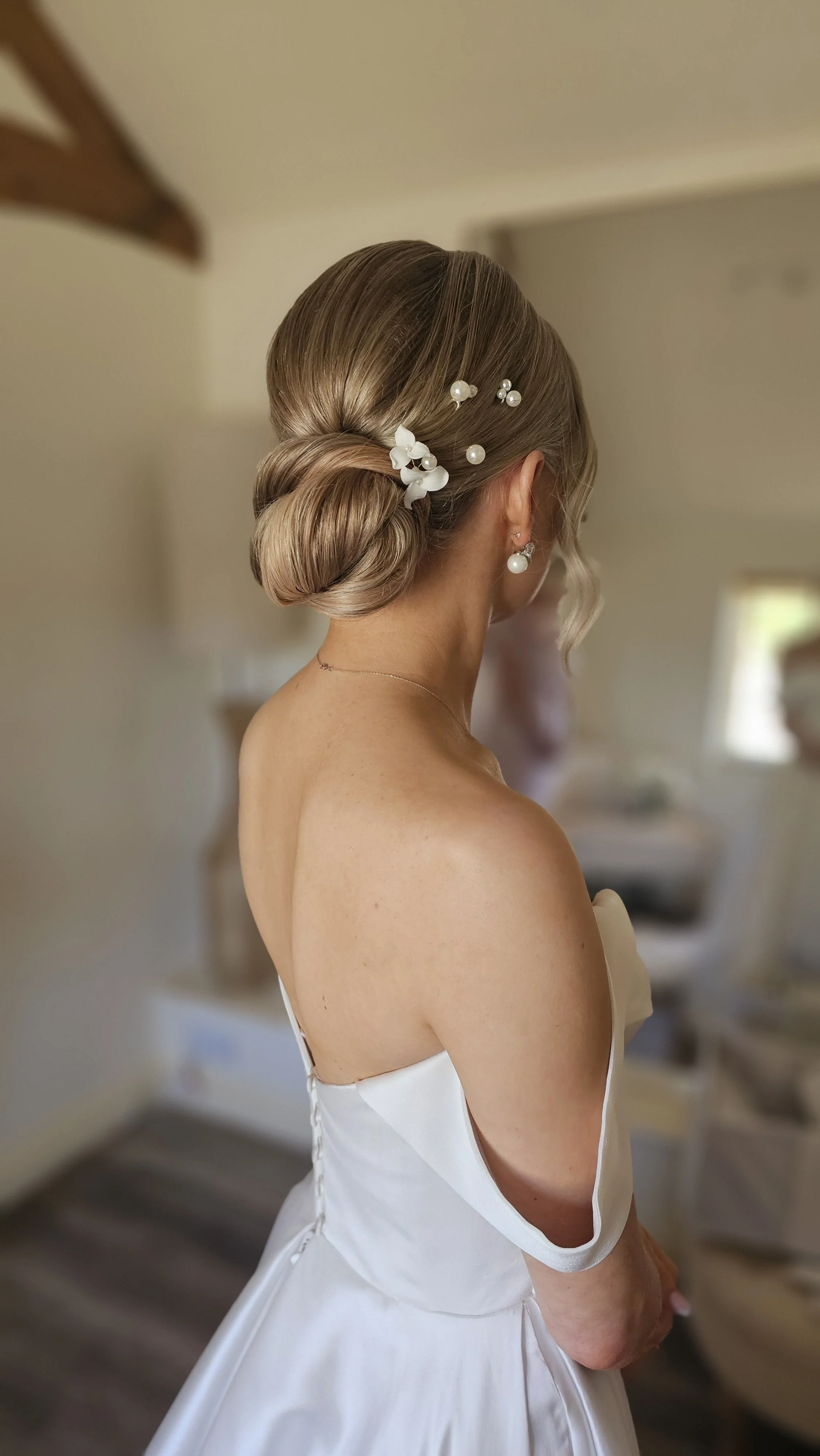 A woman with blonde hair styled in an elegant low bun, adorned with white floral and pearl hair accessories, wearing pearl earrings and a white off-shoulder dress.