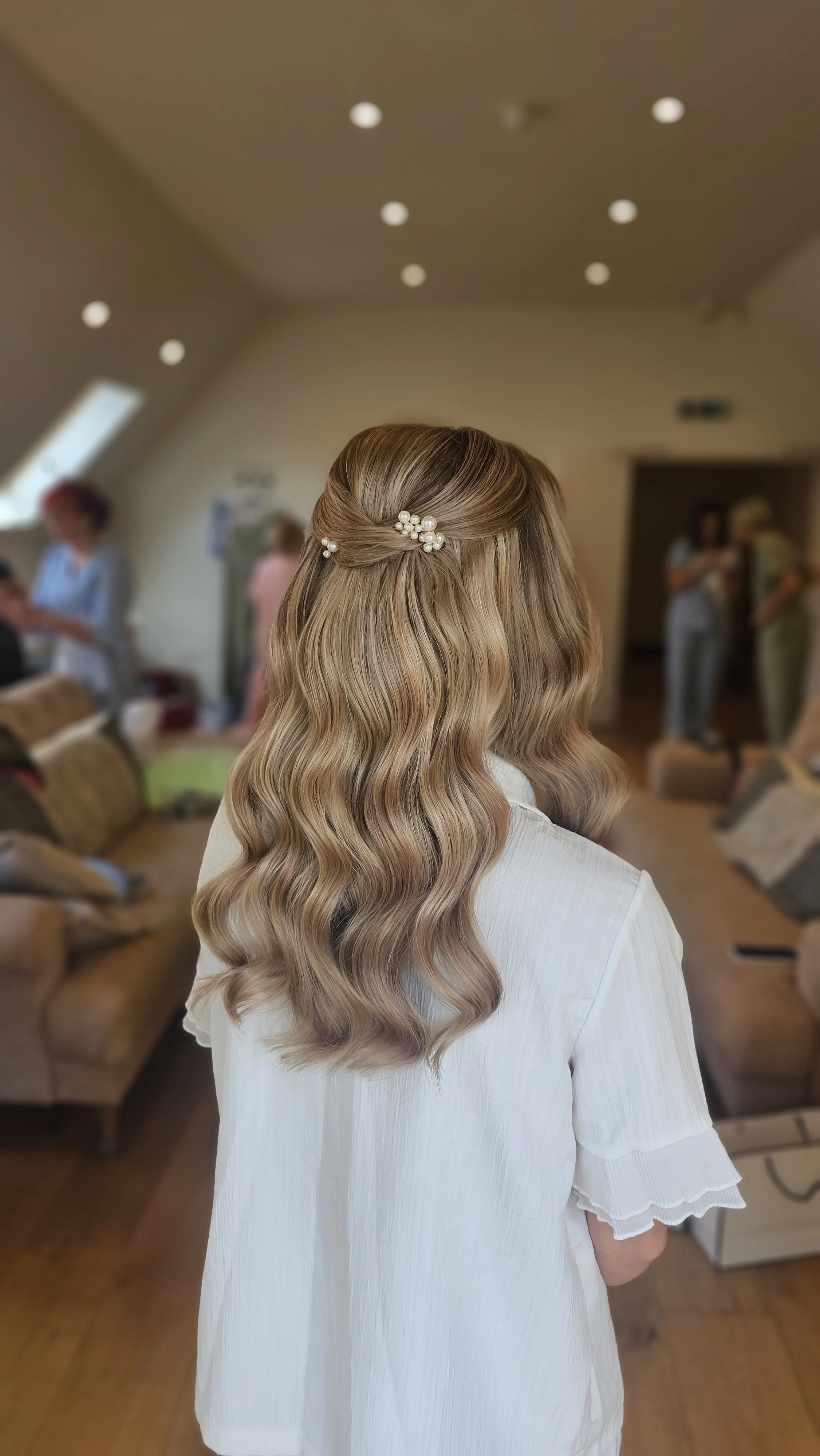 Back view of a woman with long, wavy blonde hair decorated with a pearl hair clip, wearing a white dress, in a room with blurred background of people and furniture.