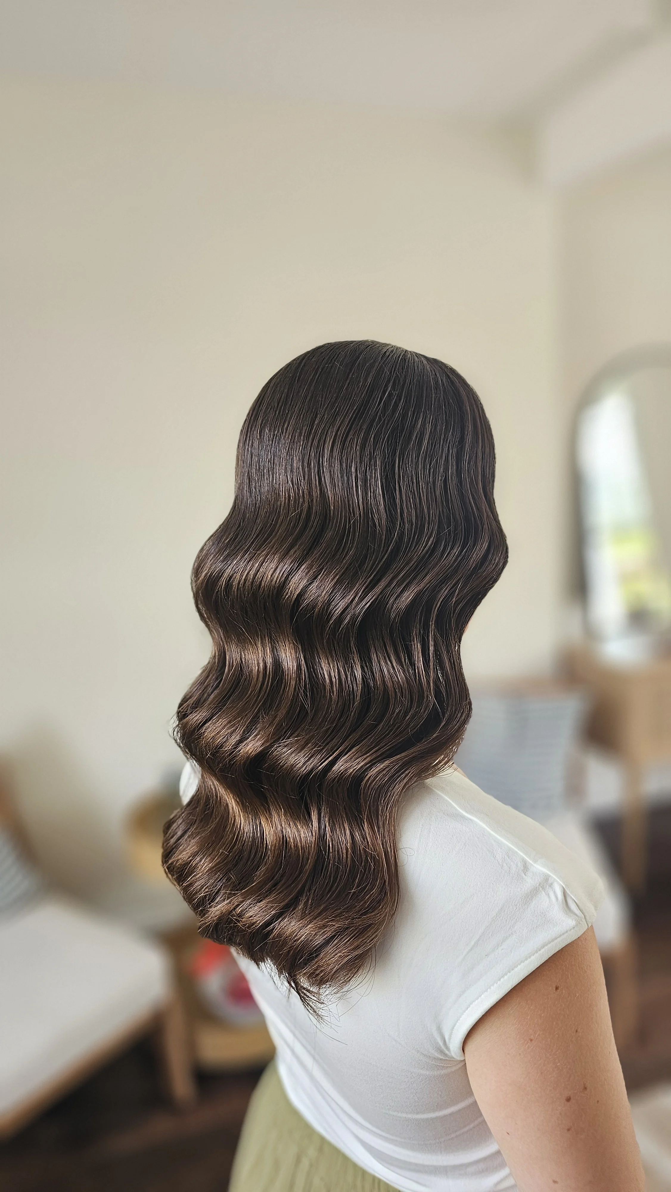Back view of a woman with medium-length, wavy, brown hair with highlights, wearing a white top, in a living room with a mirror and sofa in the background.
