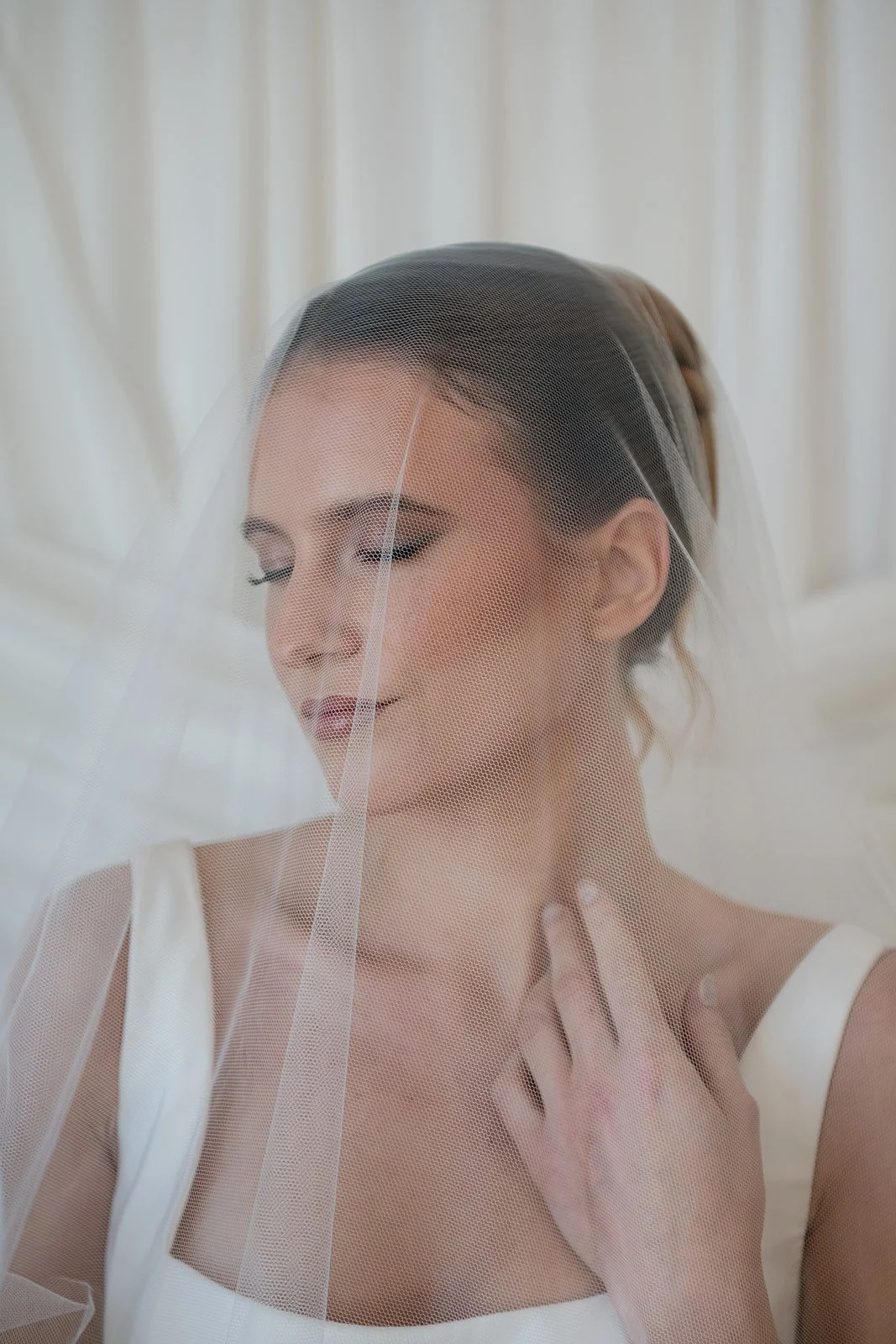 A bride with makeup, wearing a white dress and veil, rests her hand on her chest with her eyes closed, standing in front of a neutral background.