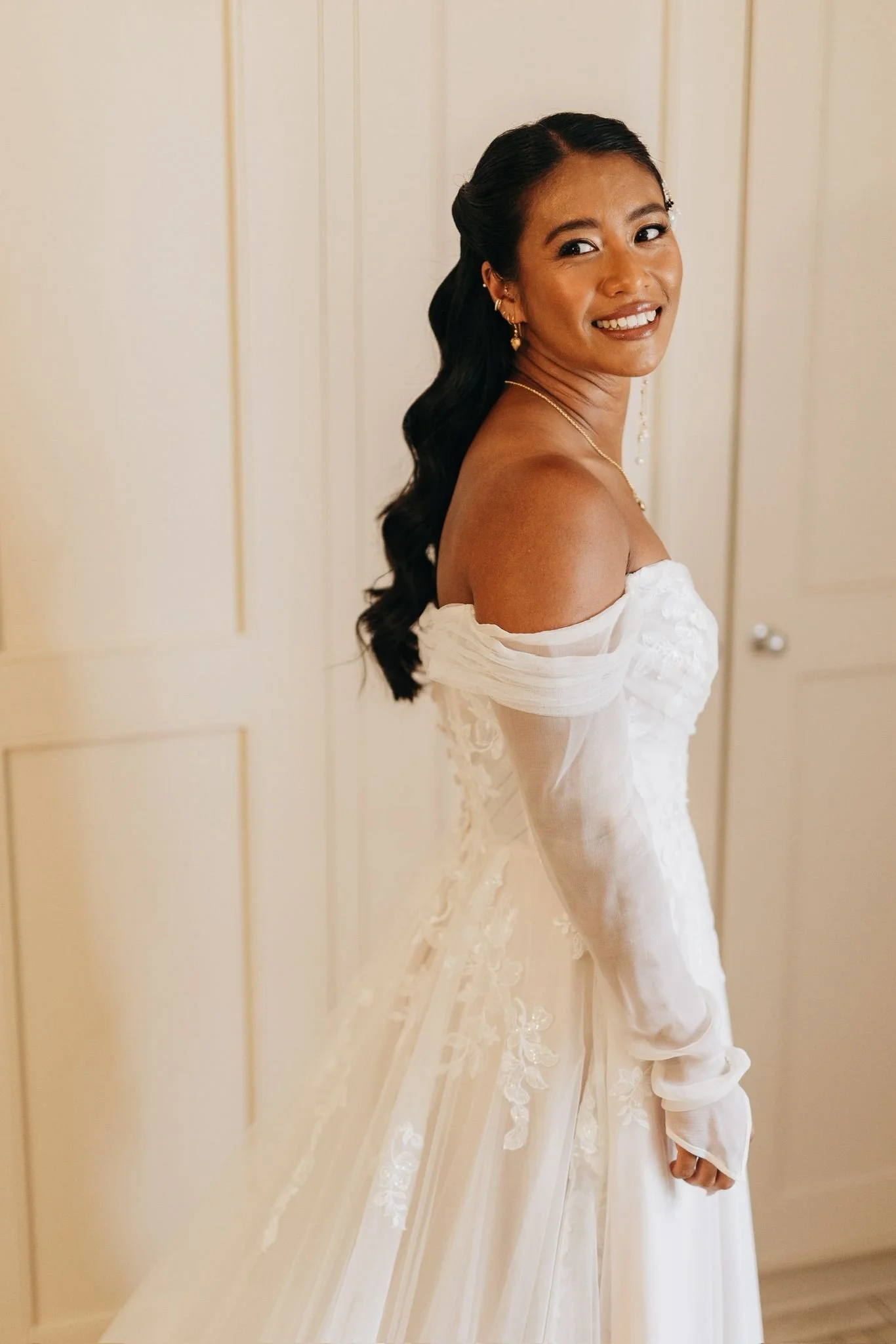 A woman in a white wedding dress with off-shoulder sleeves, smiling and looking over her shoulder, standing indoors against a cream-colored wall.