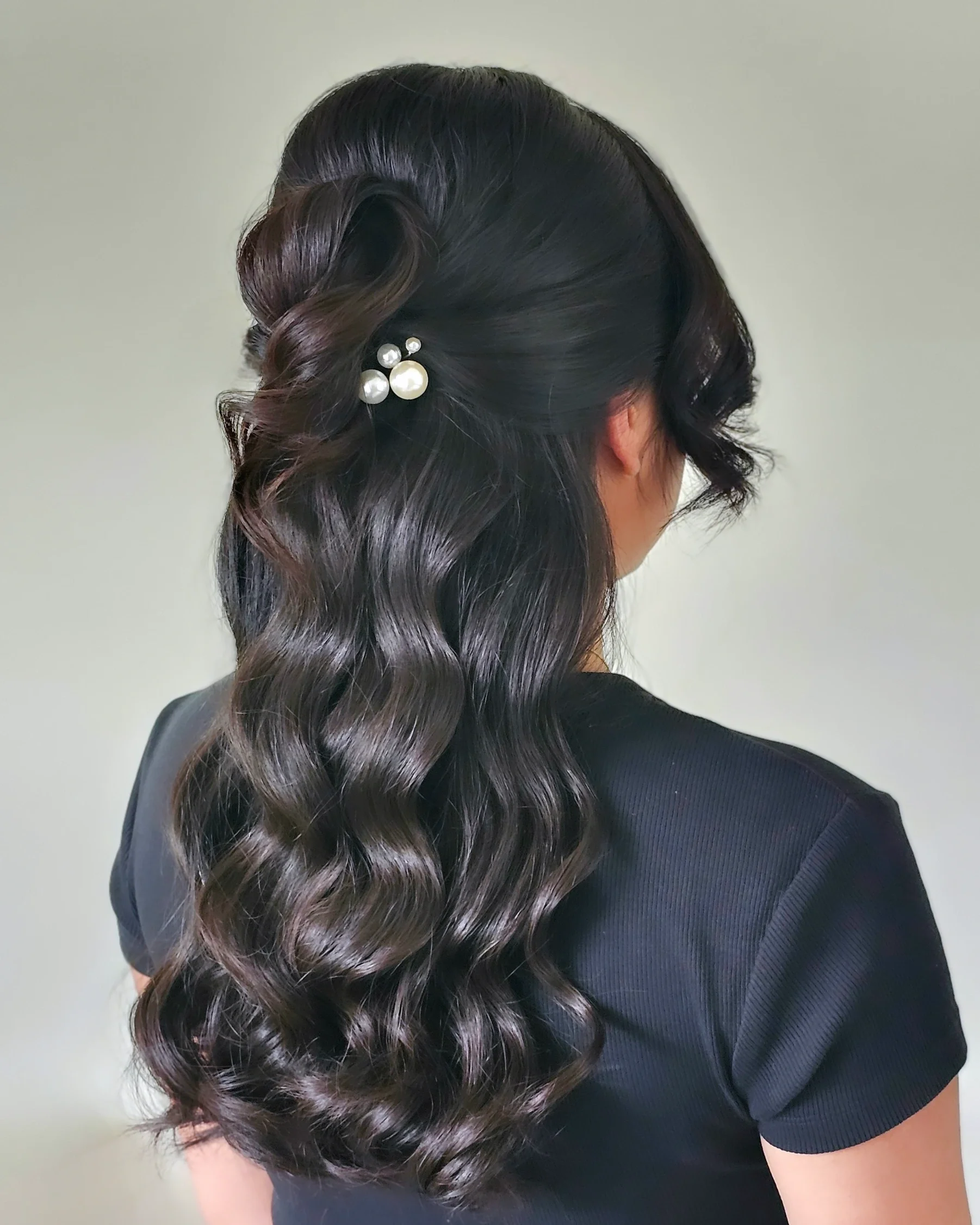 Woman with long, dark, wavy hair styled with a pearl hairpin, wearing a black shirt.