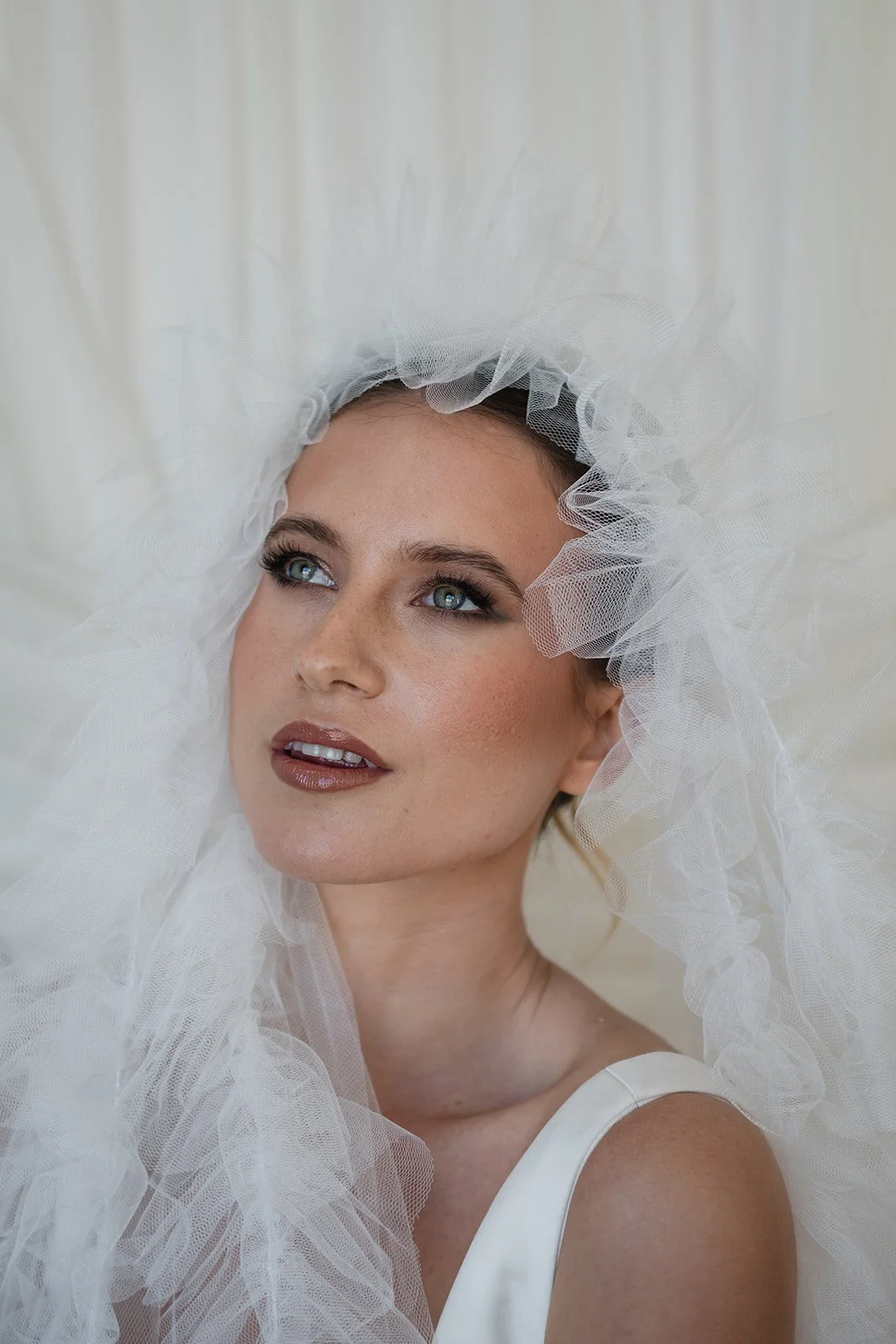 A woman with blue eyes, makeup, and long eyelashes, wearing a white dress and a tulle headpiece, looking slightly upward.