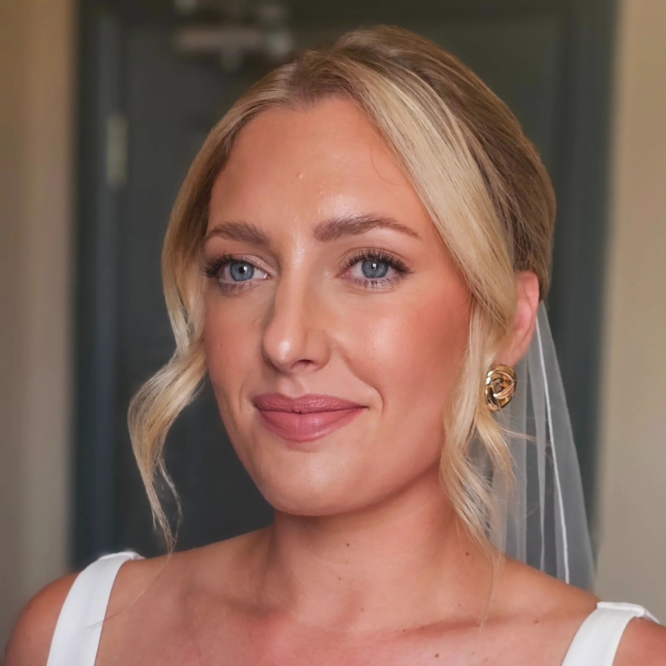 Close-up of a woman with blonde hair styled in loose waves, wearing gold earrings and a white top, smiling softly, with a background indicating an indoor setting.