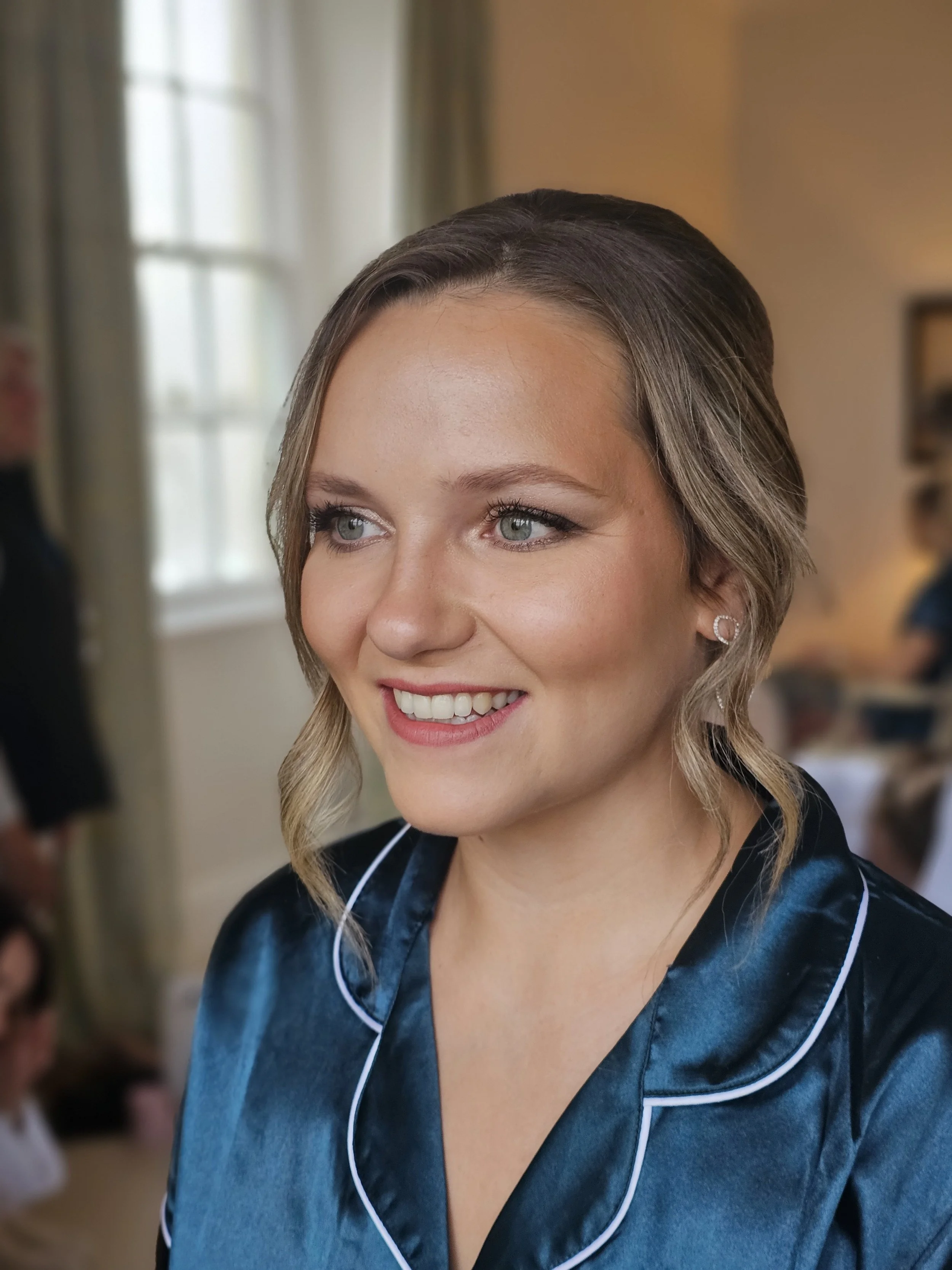 Close-up of a smiling woman with light skin, blue eyes, and wavy blonde hair, wearing blue satin pajamas with white piping, in a softly lit room with a window and blurry background.