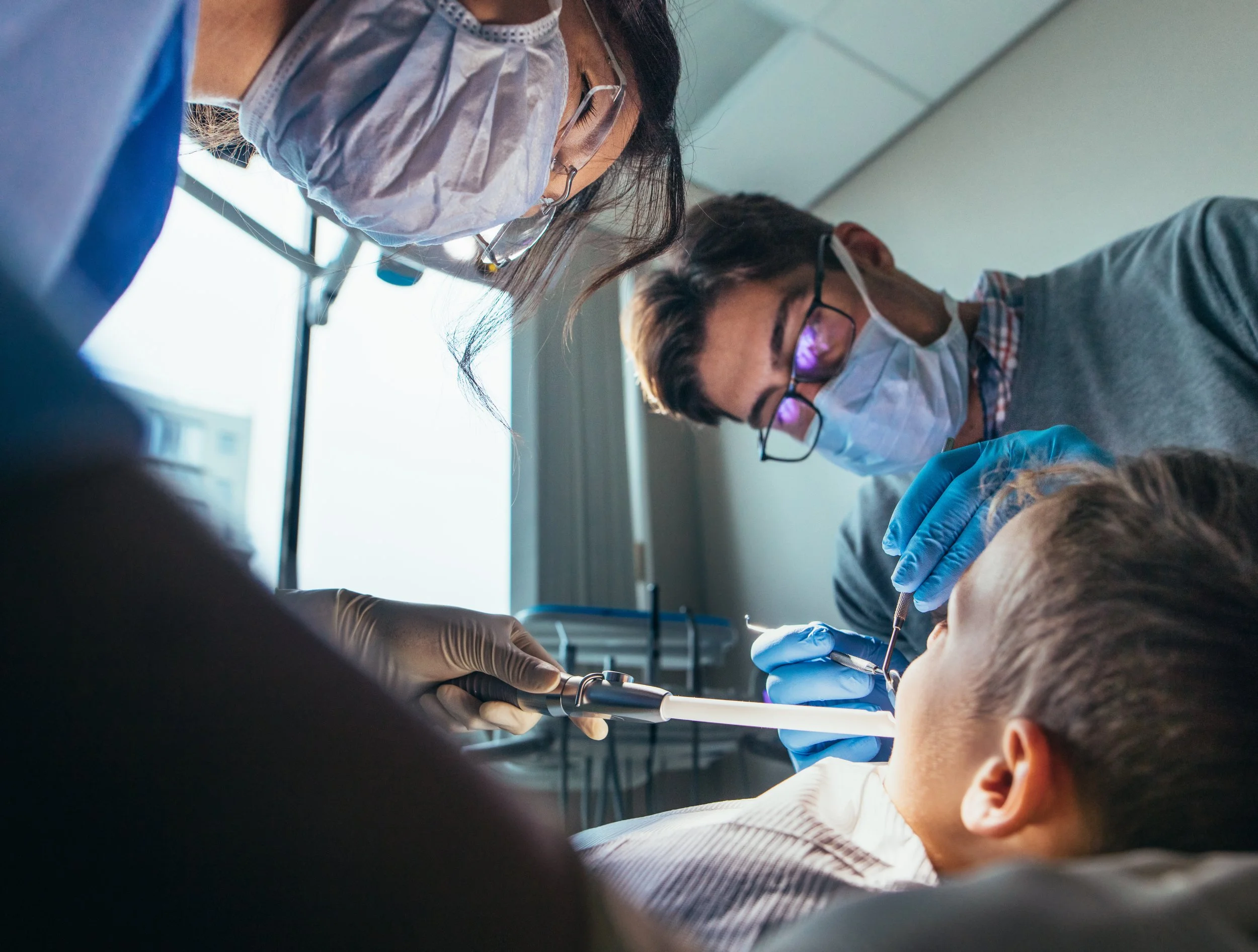 Dentist performing dental surgery on a patient with dental tools, while two dental assistants observe, all wearing face masks and gloves.