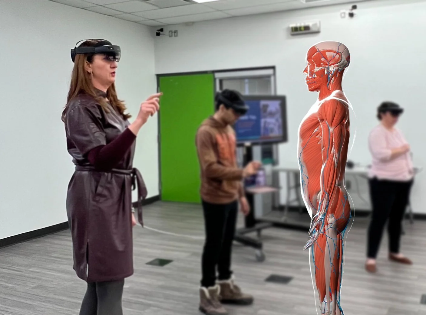 A woman wearing a virtual reality headset and a brown dress is pointing at an anatomical model of a human body with muscles and nerves, in a classroom setting with two other people also wearing VR headsets and a green door in the background.