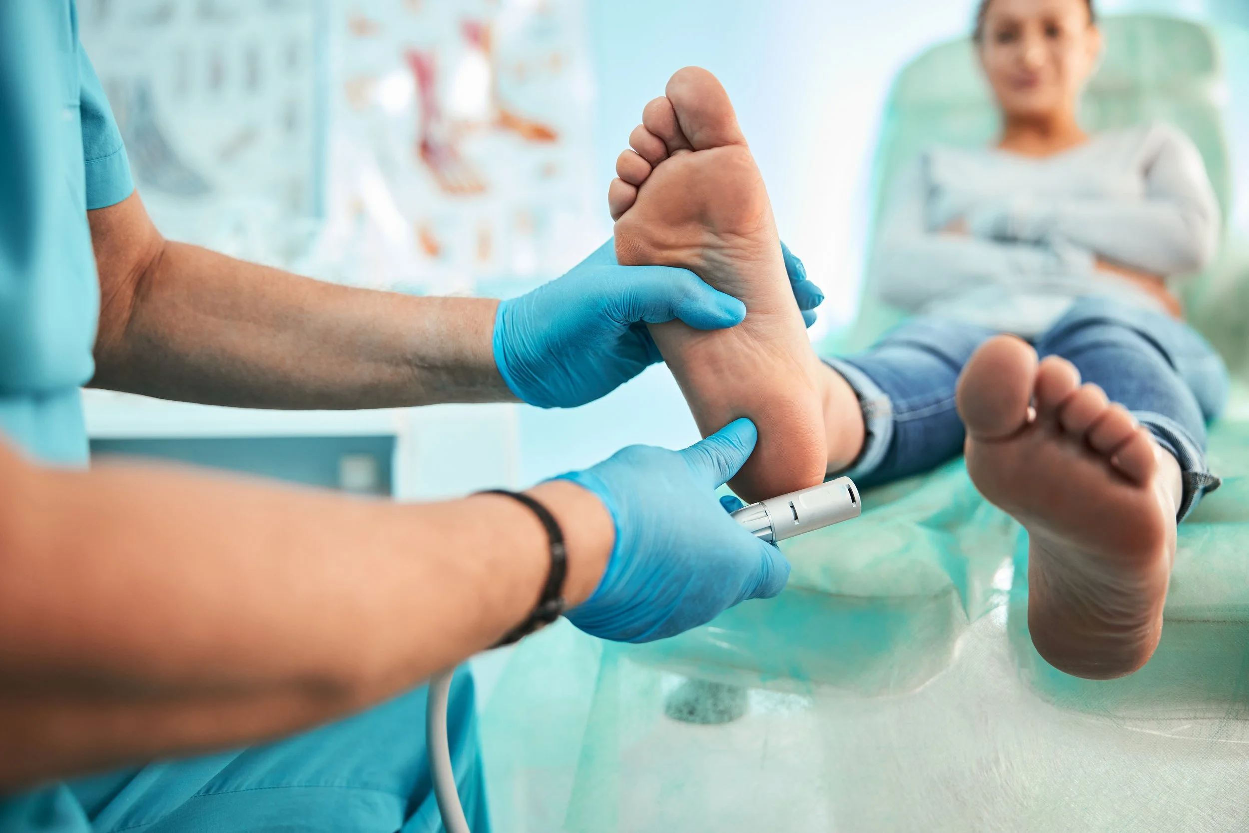 Medical professional wearing blue gloves examining a child's foot with a medical tool in a clinic setting, with the child lying on a medical bed.