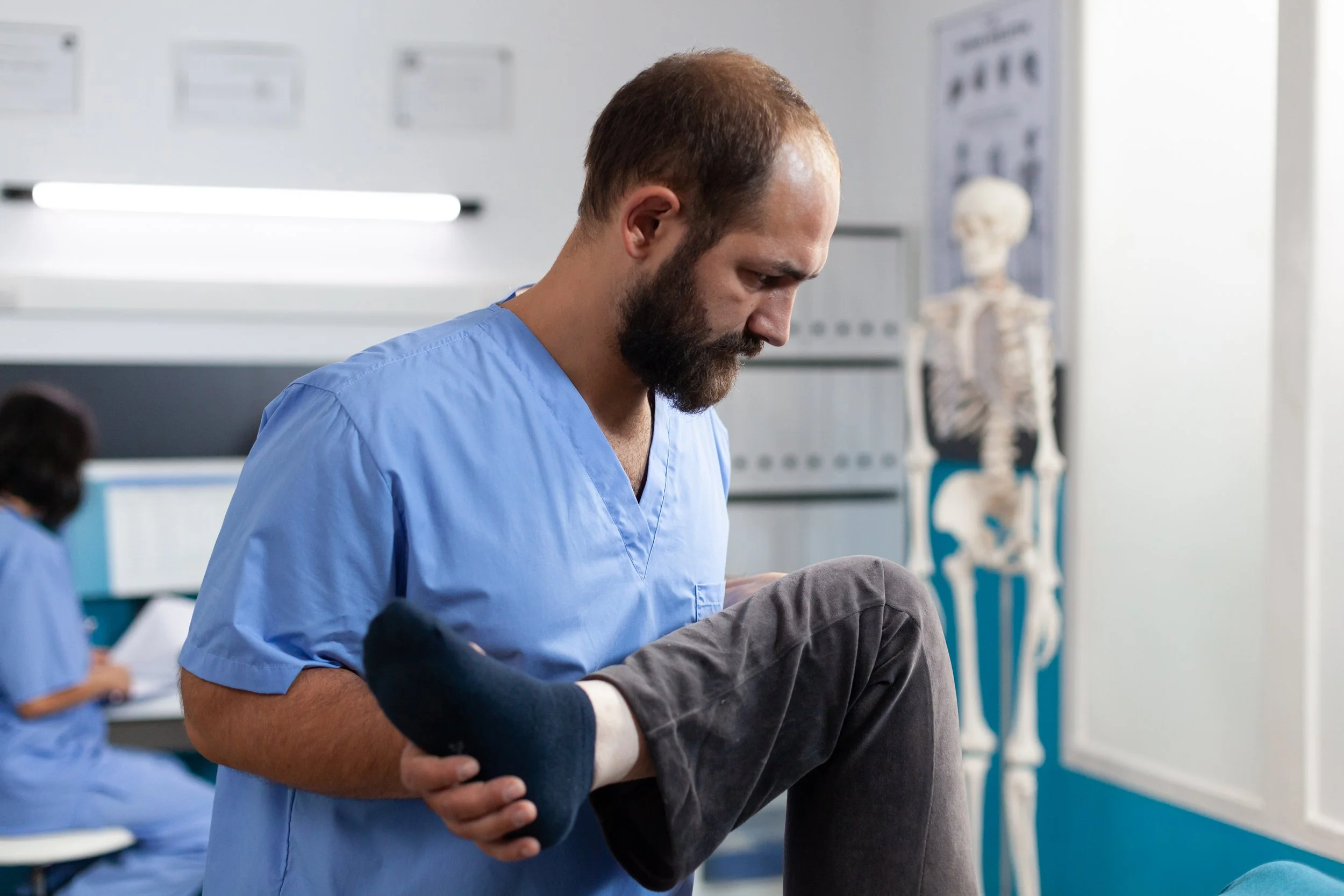 A male healthcare professional, wearing blue scrubs, is examining a patient's foot in a medical setting. An anatomical skeleton model and another healthcare worker are visible in the background.