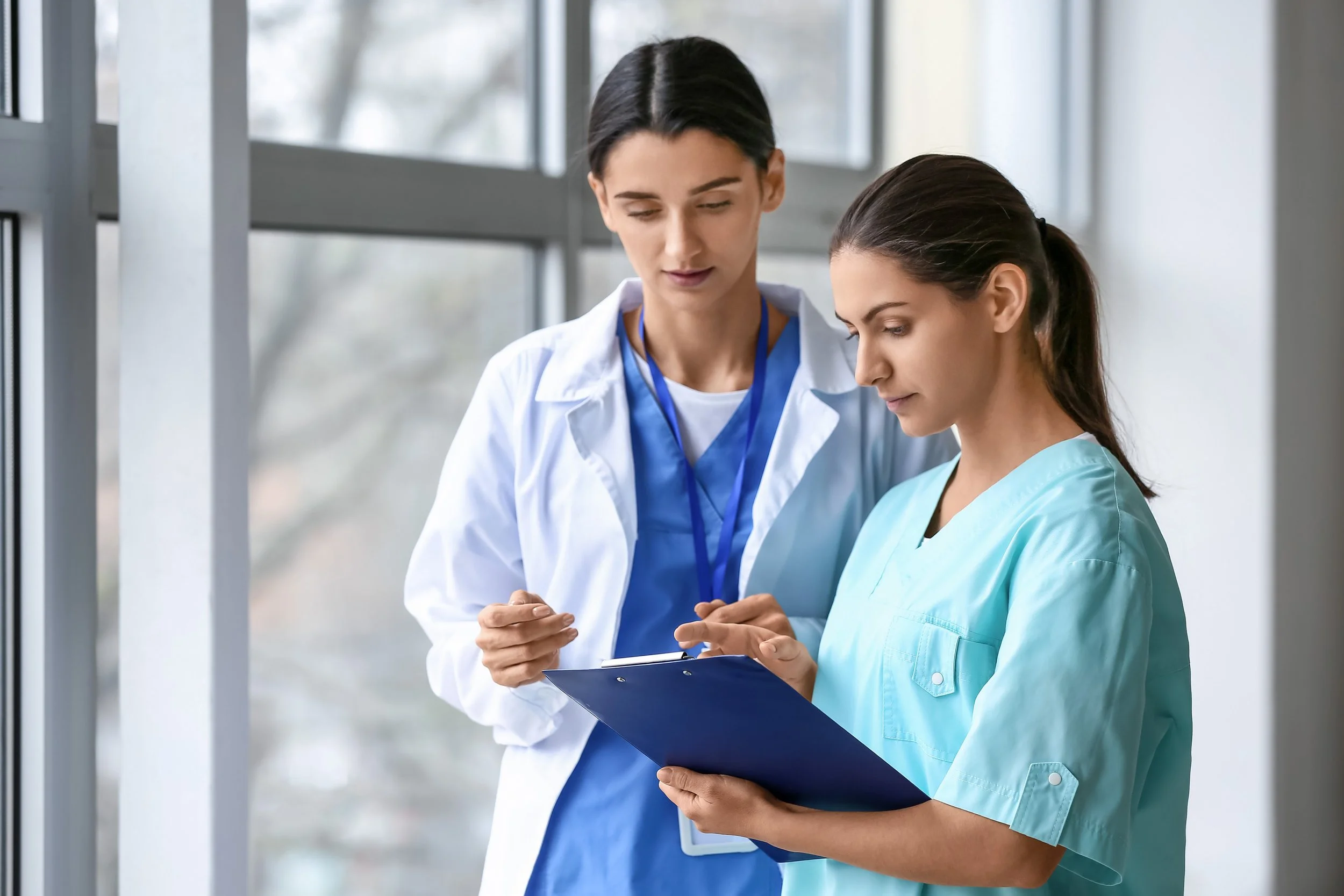 Two healthcare professionals, a woman in a white coat and a woman in light blue scrubs, reviewing documents together in a medical facility with large windows.