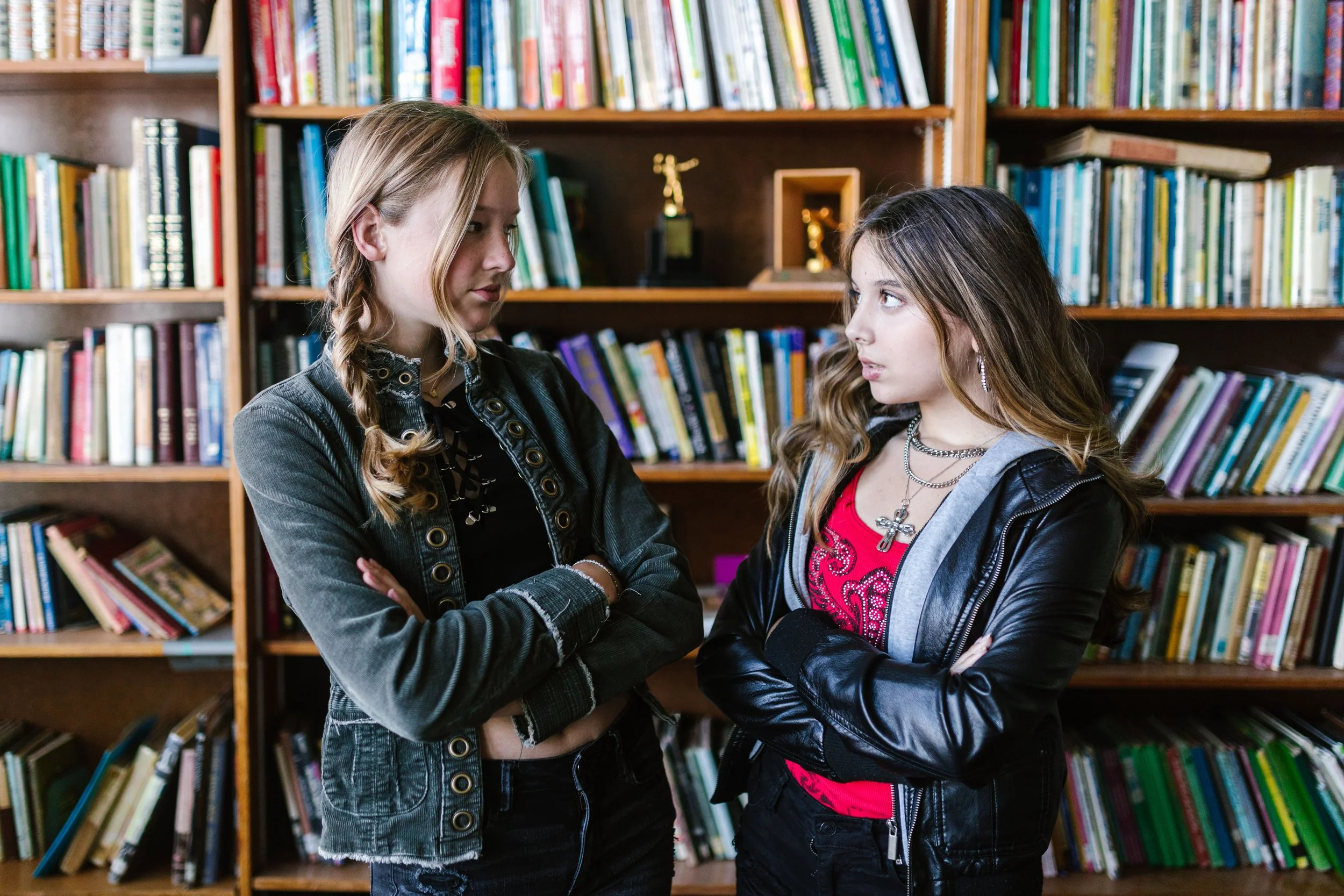 Two teen girls stand with arms crossed, exchanging a tense look in a library. What does teen conflict really look like between former friends? Teen therapy in San Ramon, CA can help kids work through these hard feelings.