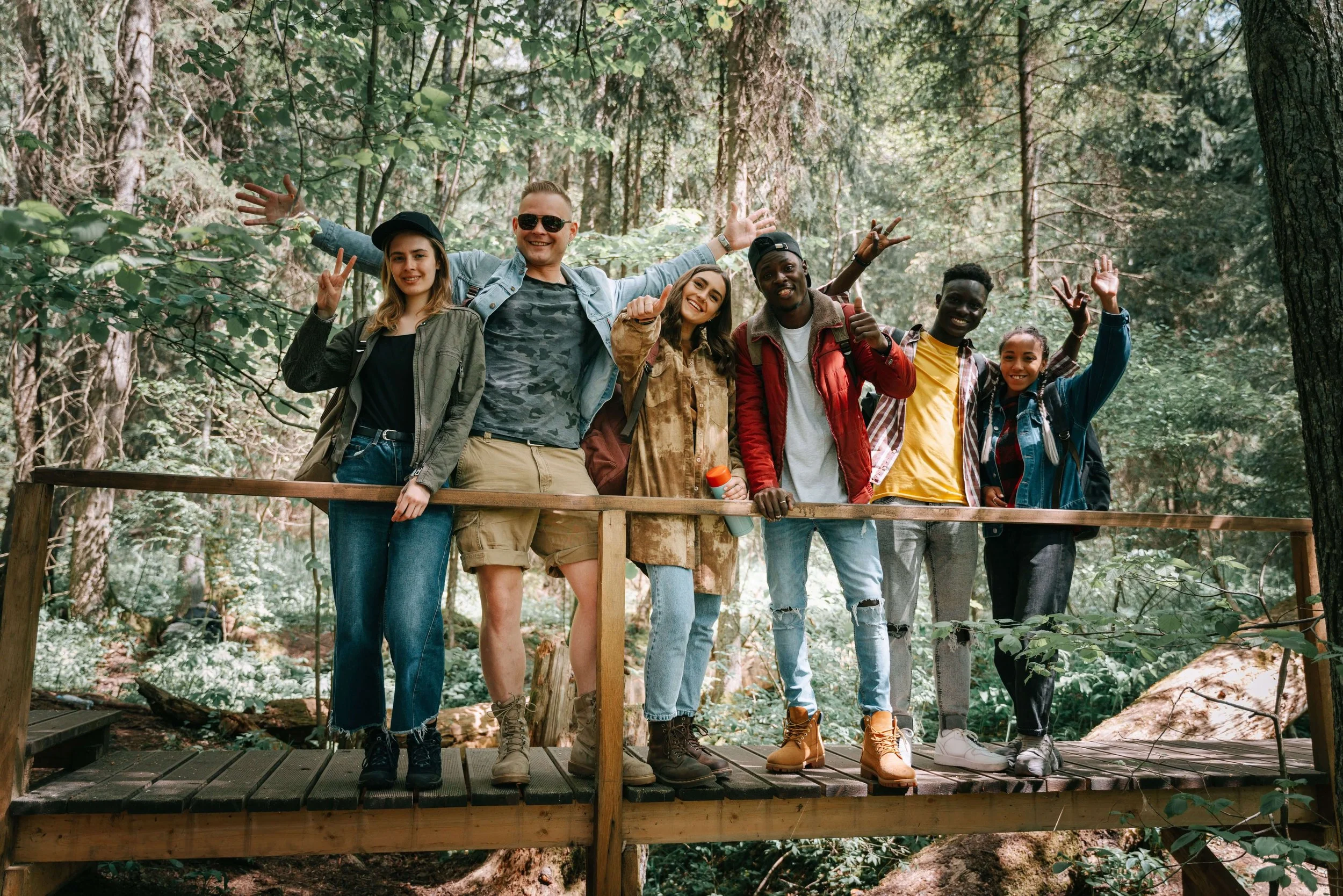 Six friends smile and pose together on a wooden bridge surrounded by trees, enjoying time outdoors.