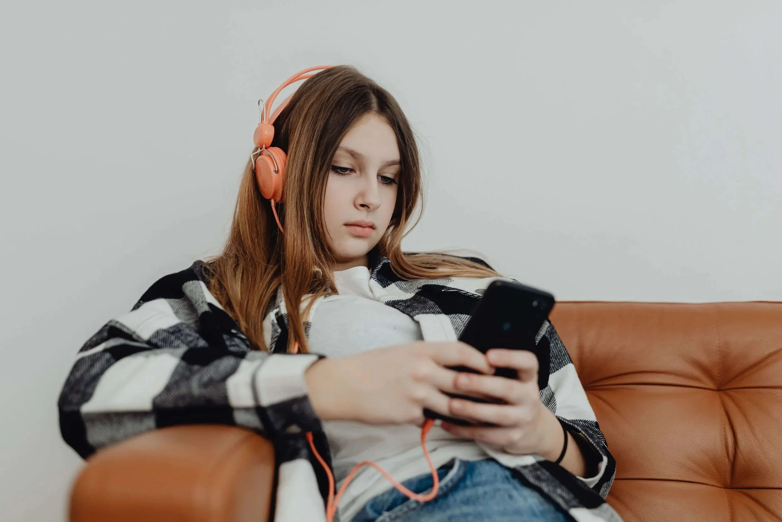 A teenage girl sitting alone on a sofa while listening to headphones attached to a smartphone.