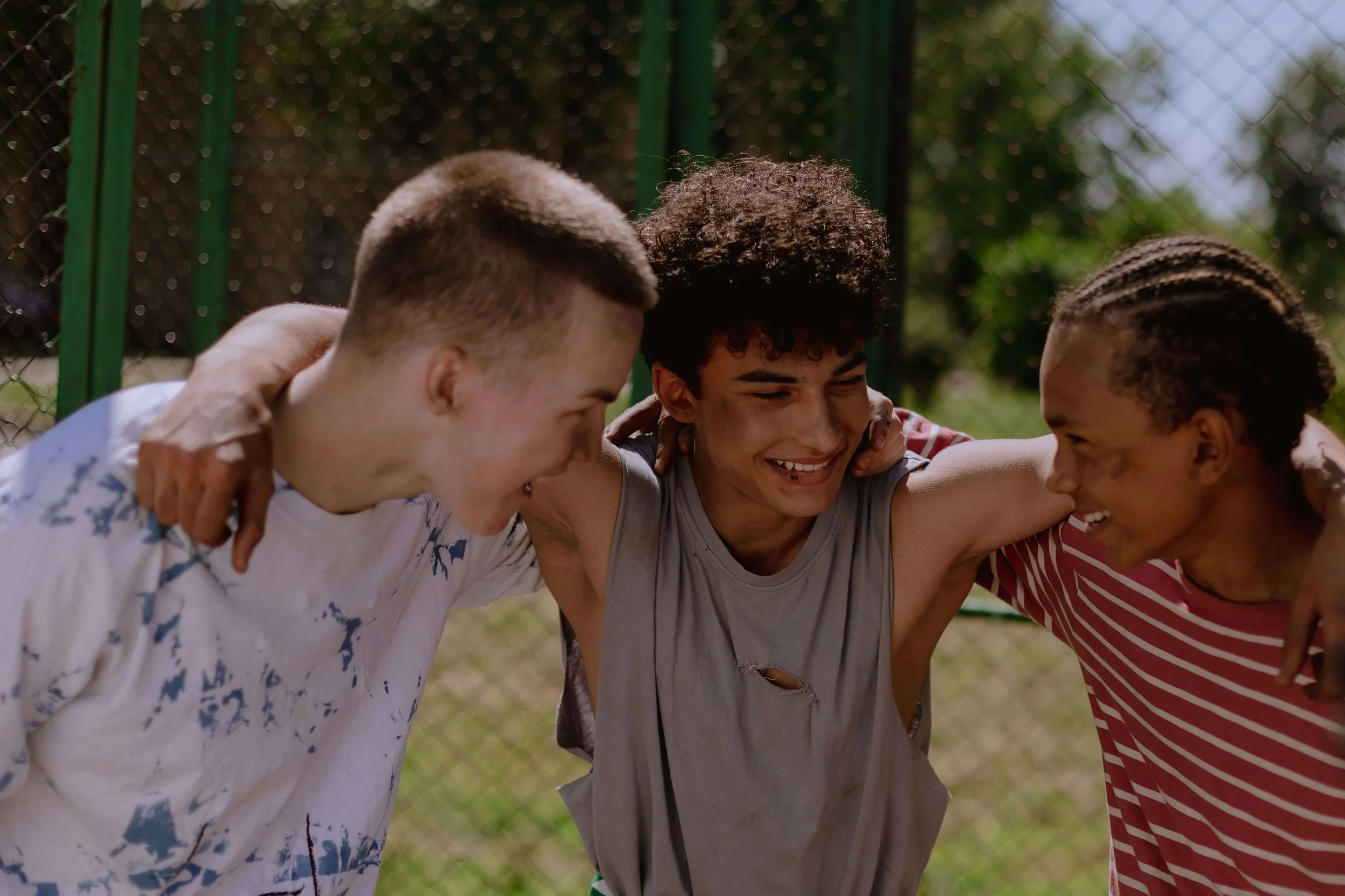 Three teenage boys smile and laugh together outdoors with their arms around each other's shoulders.