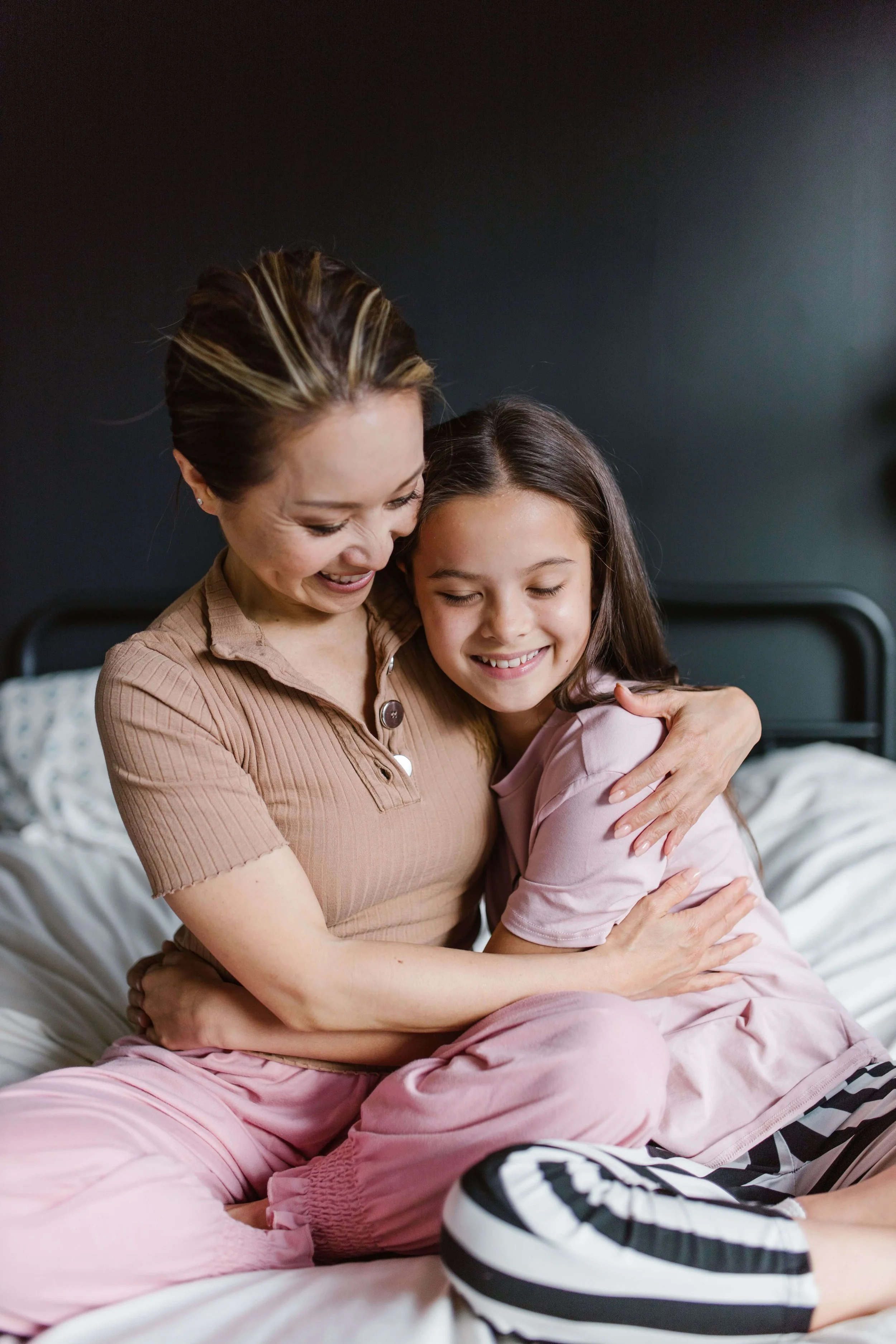 Woman and young girl smiling and hugging while sitting together on a bed, sharing a warm moment of connection.