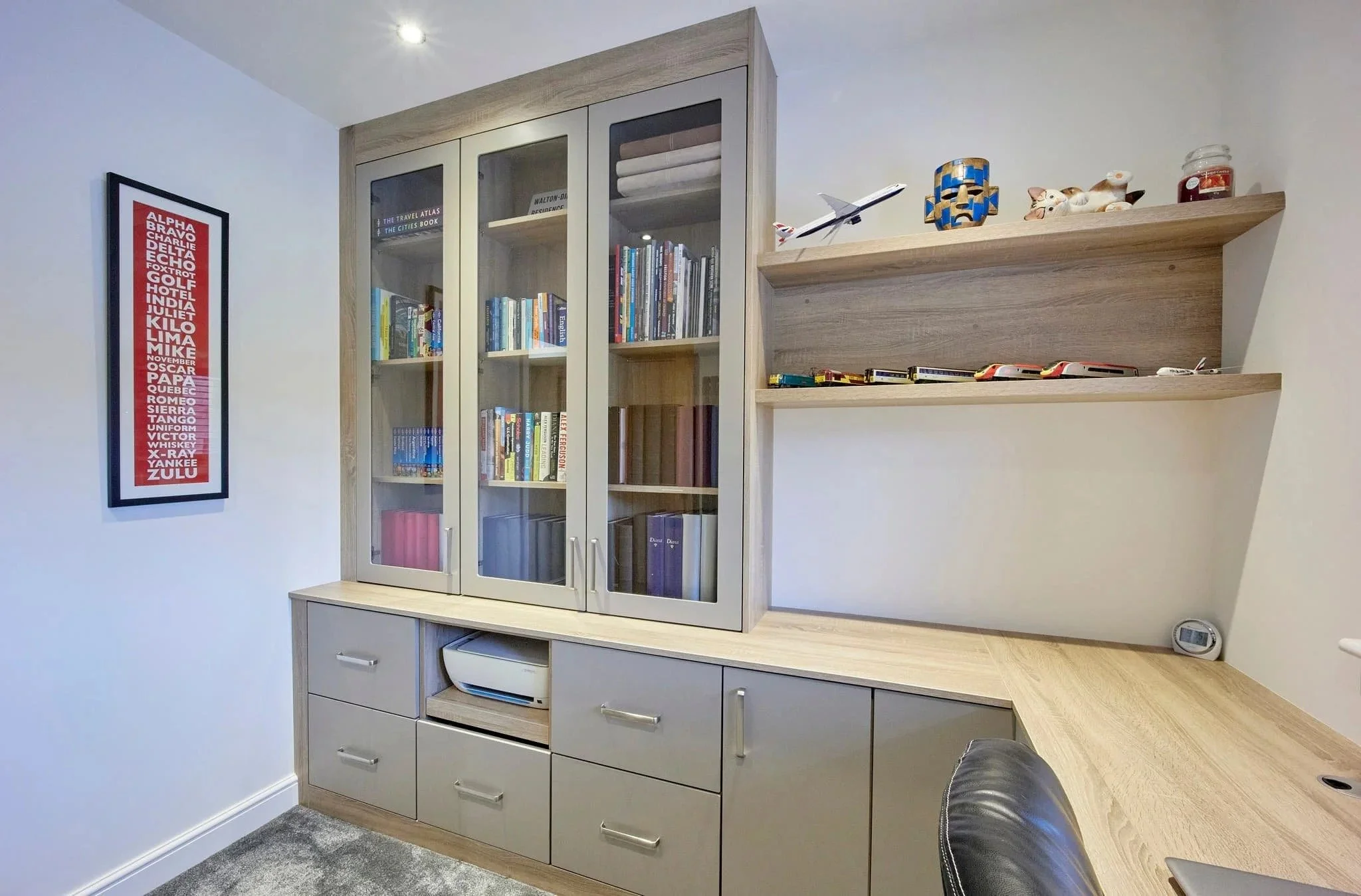 Modern fitted home office furniture with grey cabinets, glass bookcase storage, wooden desk, and floating shelves.