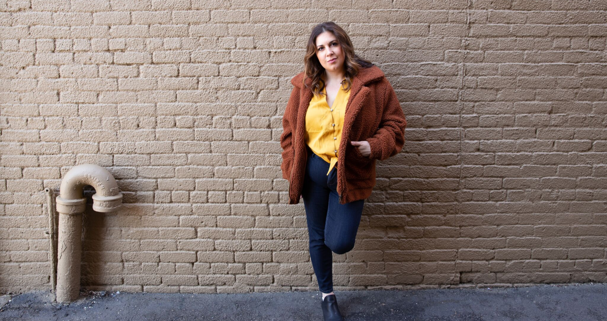 Woman, Noelle Rhodes, in yellow shirt and brown coat leaning against a brick wall.