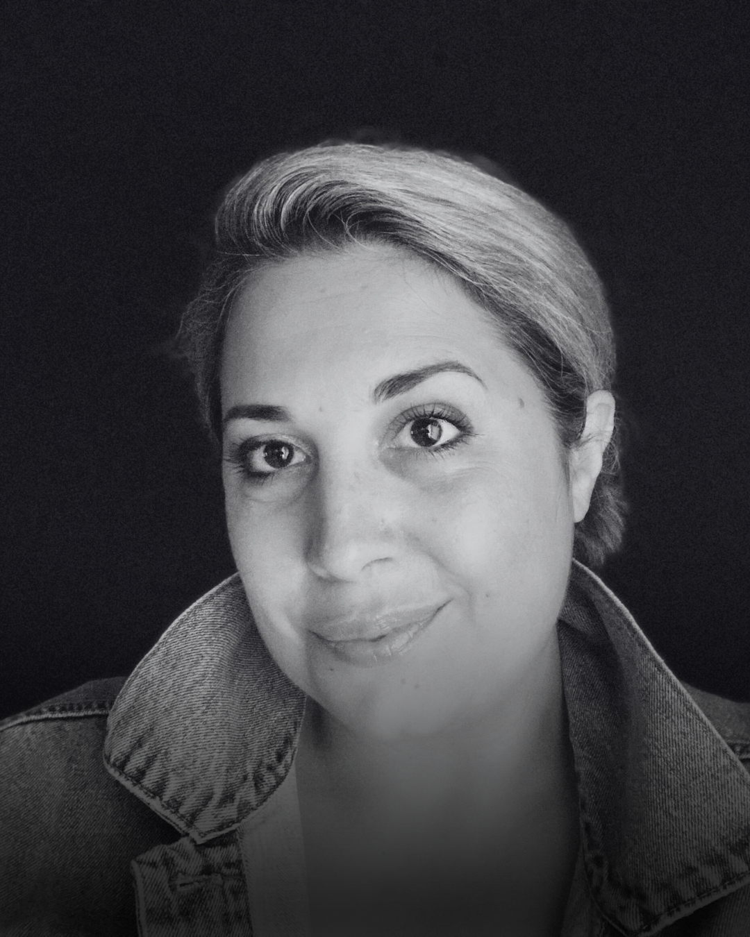 Black and white portrait of Noelle Rhodes with hair pulled back, wearing a denim jacket, smiling softly with a neutral dark background.