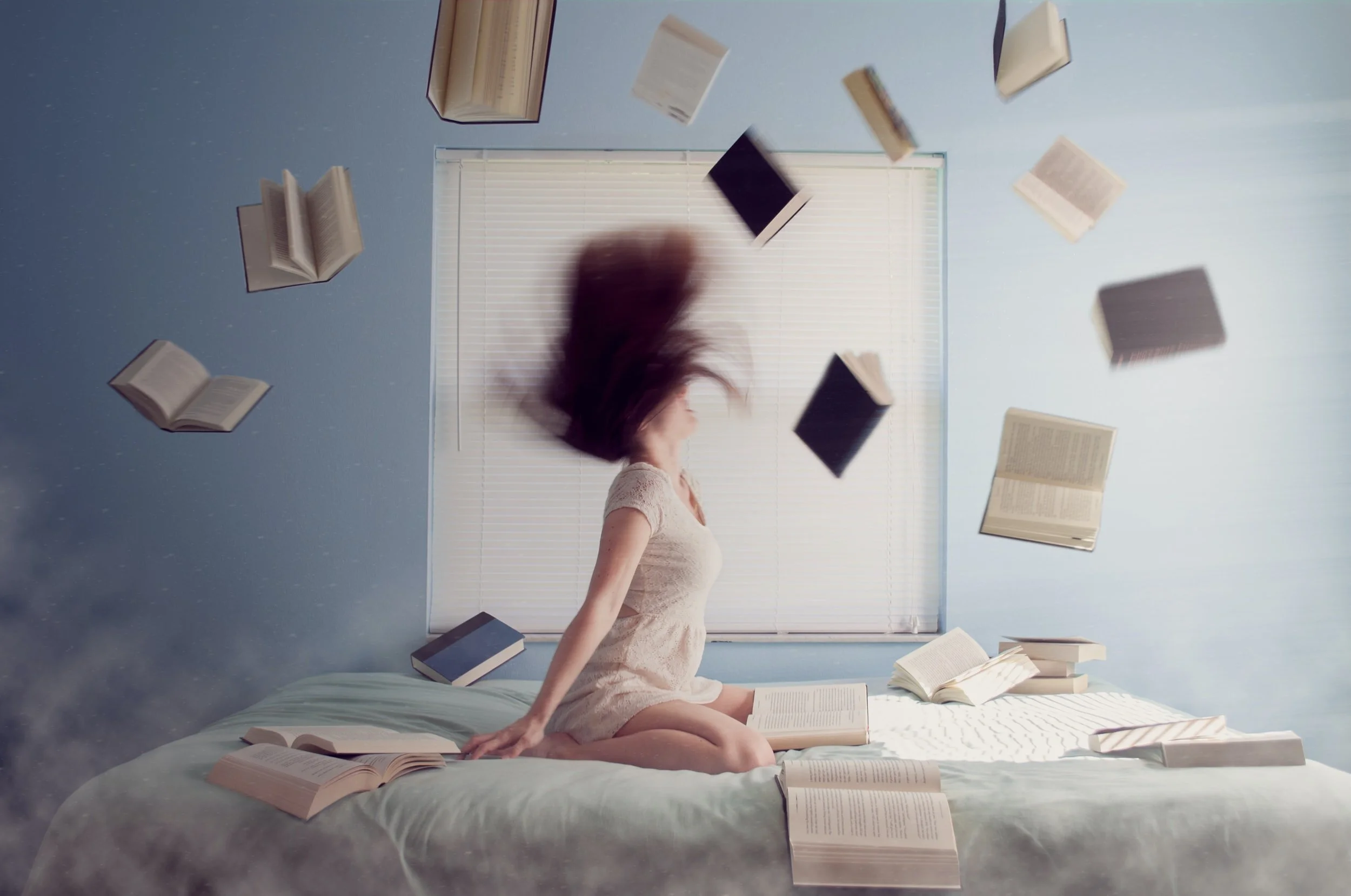 Woman tosses books in the air while sitting on top of a light green comforter in a blue bedroom. If you're feeling overwhelmed by anger or anxiety, try therapy in NYC here.