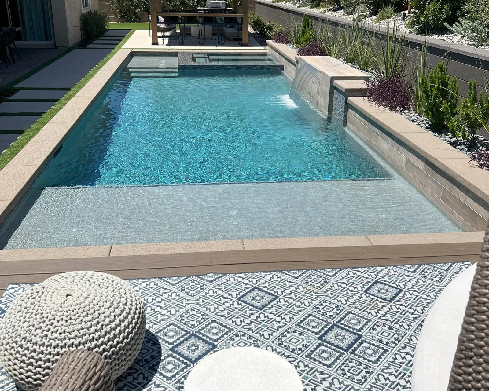 A modern backyard swimming pool, with a  large tanning ledge, a  shear decent water feature, and an elevated seating area.