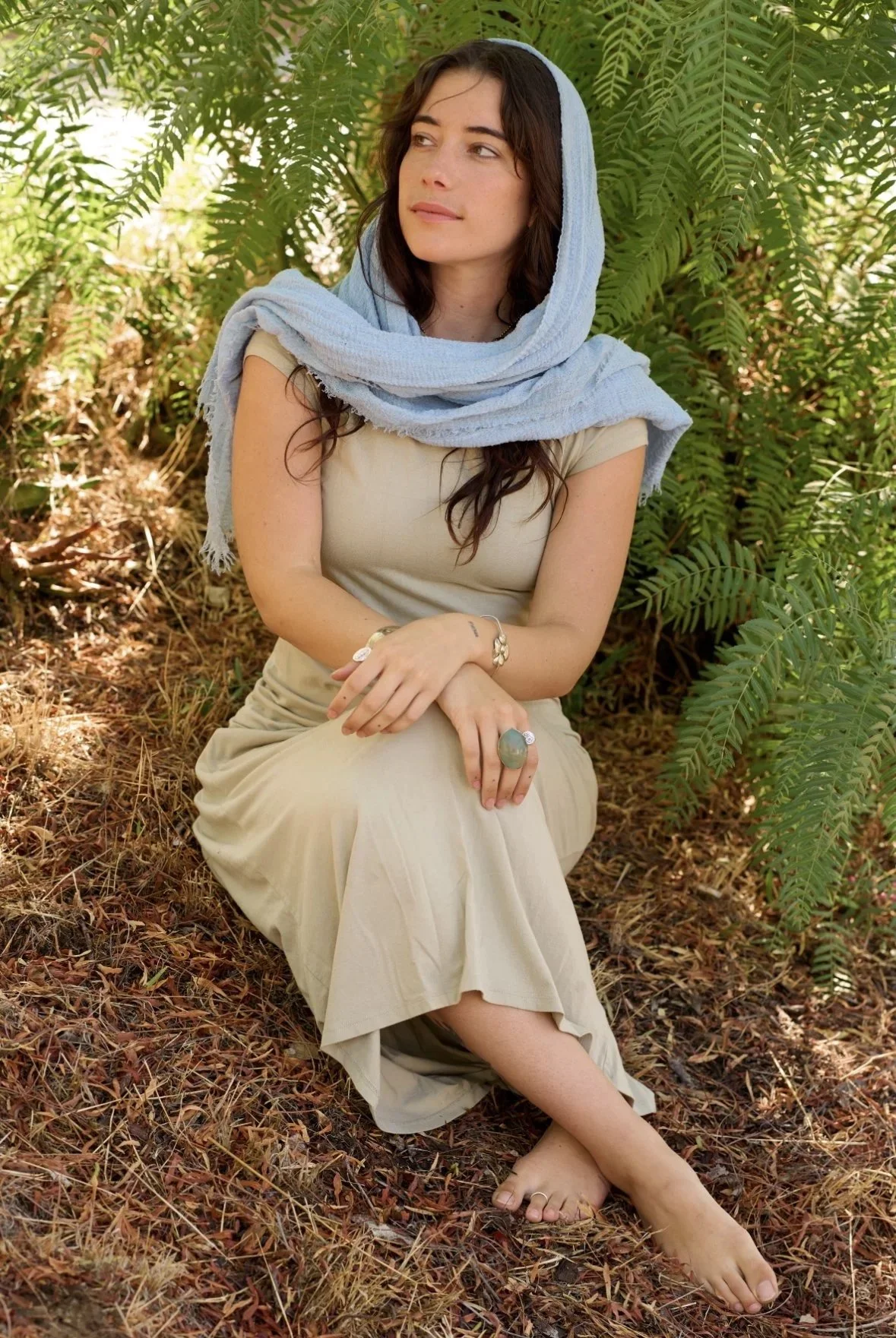 A woman with dark hair sitting outdoors among green ferns with a light blue scarf over her head and shoulders, wearing a beige dress and jewelry.