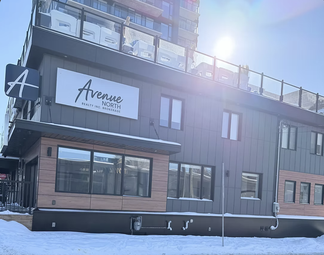 Exterior of a modern, multi-story building with a sign that reads 'Avenue North Realty Inc. Brokerage.' The building has large windows, gray and wood panel siding, and a rooftop terrace with glass railing. There is snow on the ground, and the sun is shining brightly.