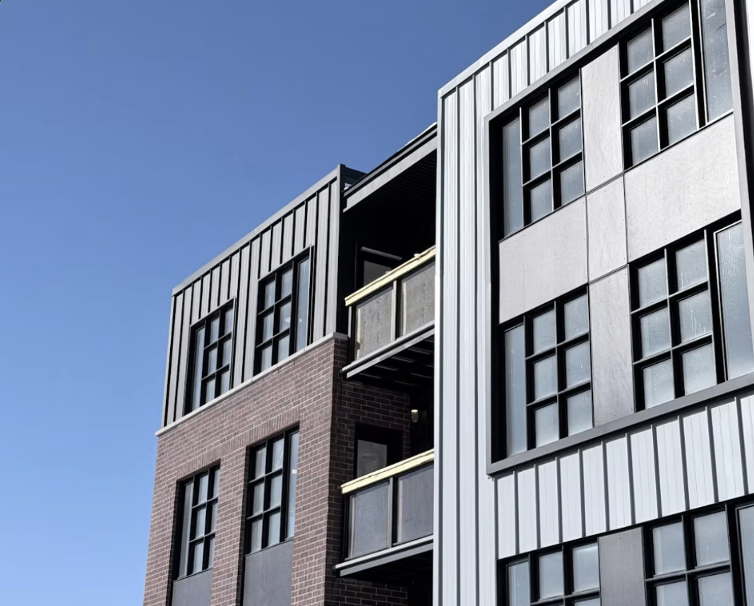 Close-up view of modern multi-story residential building with brick and metal exterior, multiple windows, and balconies under a clear blue sky.