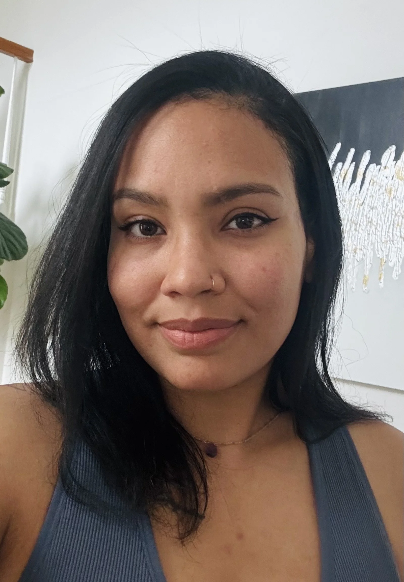 A young woman with black hair, wearing a blue tank top, a small nose piercing, and a necklace, smiling indoors with a white wall, a flower, and a modern art piece in the background.