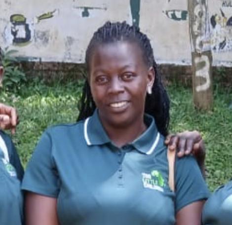 Smiling woman in green polo shirt with 'Too Little Children' logo outdoors in a lush, green landscape with trees and bushes.