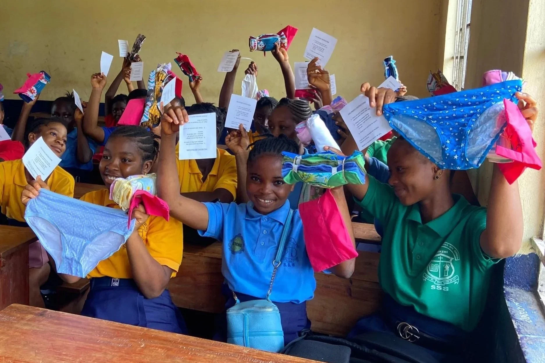 Schoolchildren in colorful uniforms celebrating in a classroom, holding up reusable sanitary pads and underwear, smiling and appearing joyful.