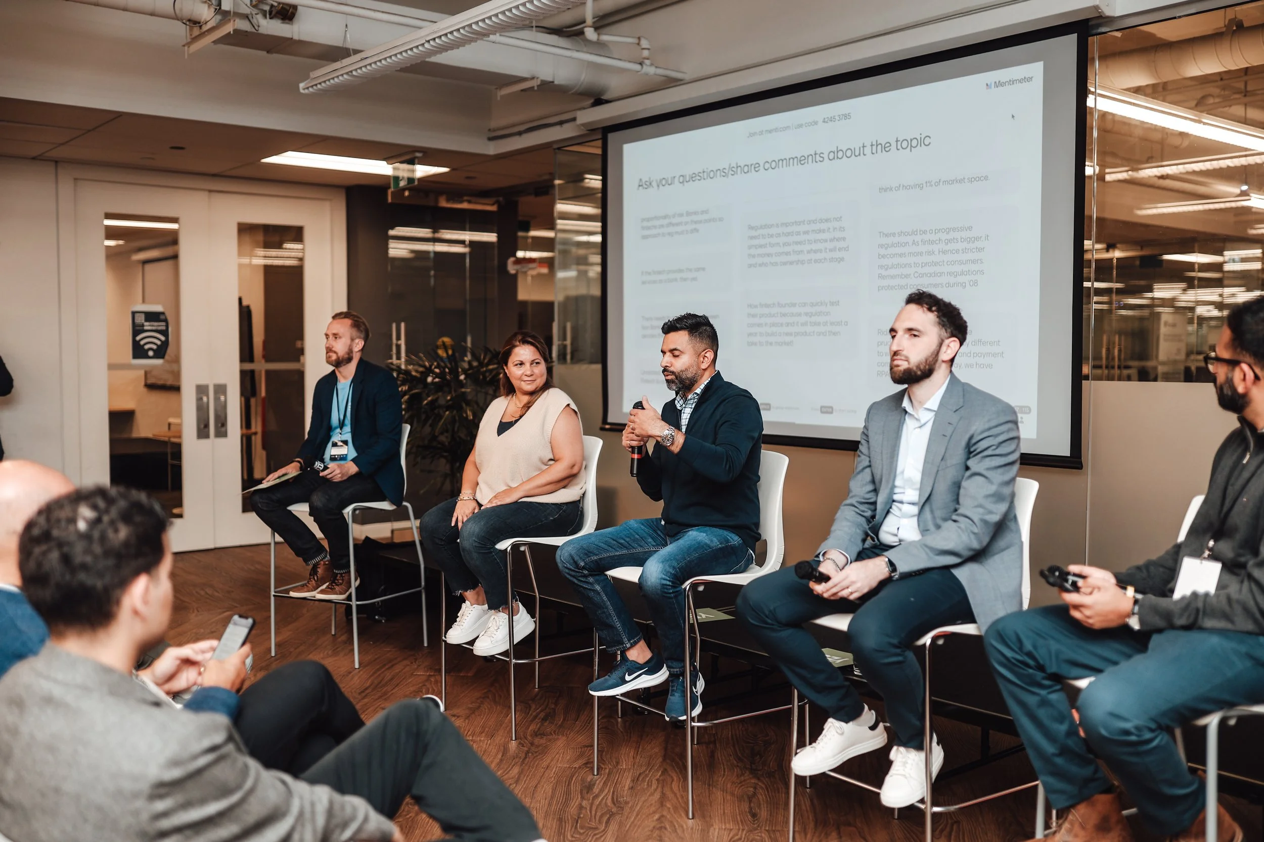 Panel of five people sitting on stools during a discussion in a conference room, with a large screen displaying a presentation behind them. The audience in front is watching, with some members using their phones.