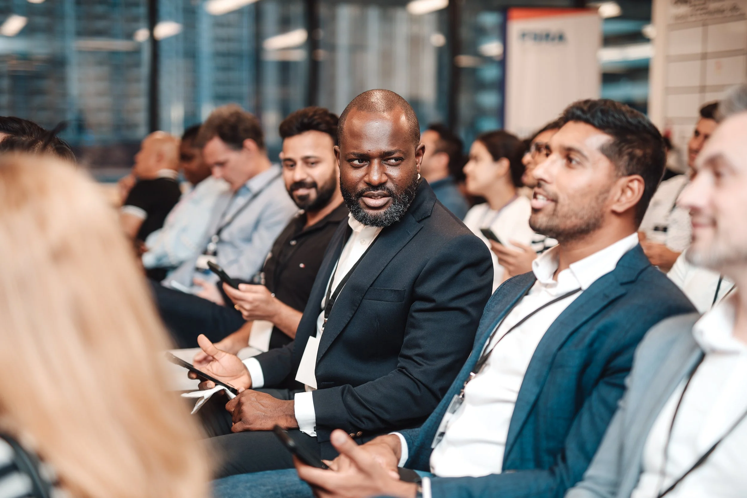 Business conference or seminar with diverse attendees engaged, some using smartphones, seated in a modern indoor setting.