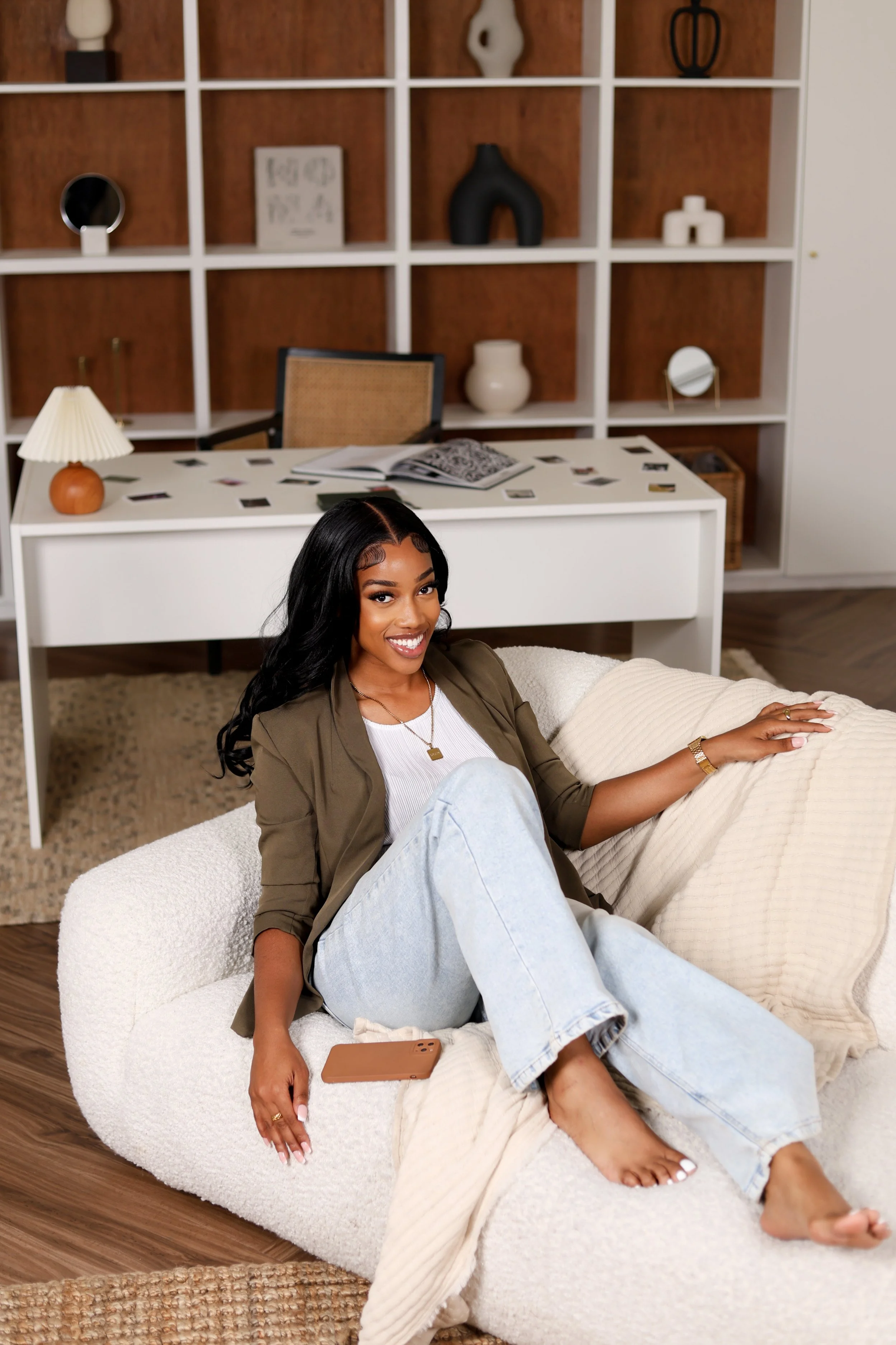 Smiling woman with long black hair, casual attire, sitting barefoot on a cozy beige sofa in a modern living room, with a white throw blanket, a smartphone nearby, and a white textured pillow, behind a white desk with a lamp and open magazine, in fron