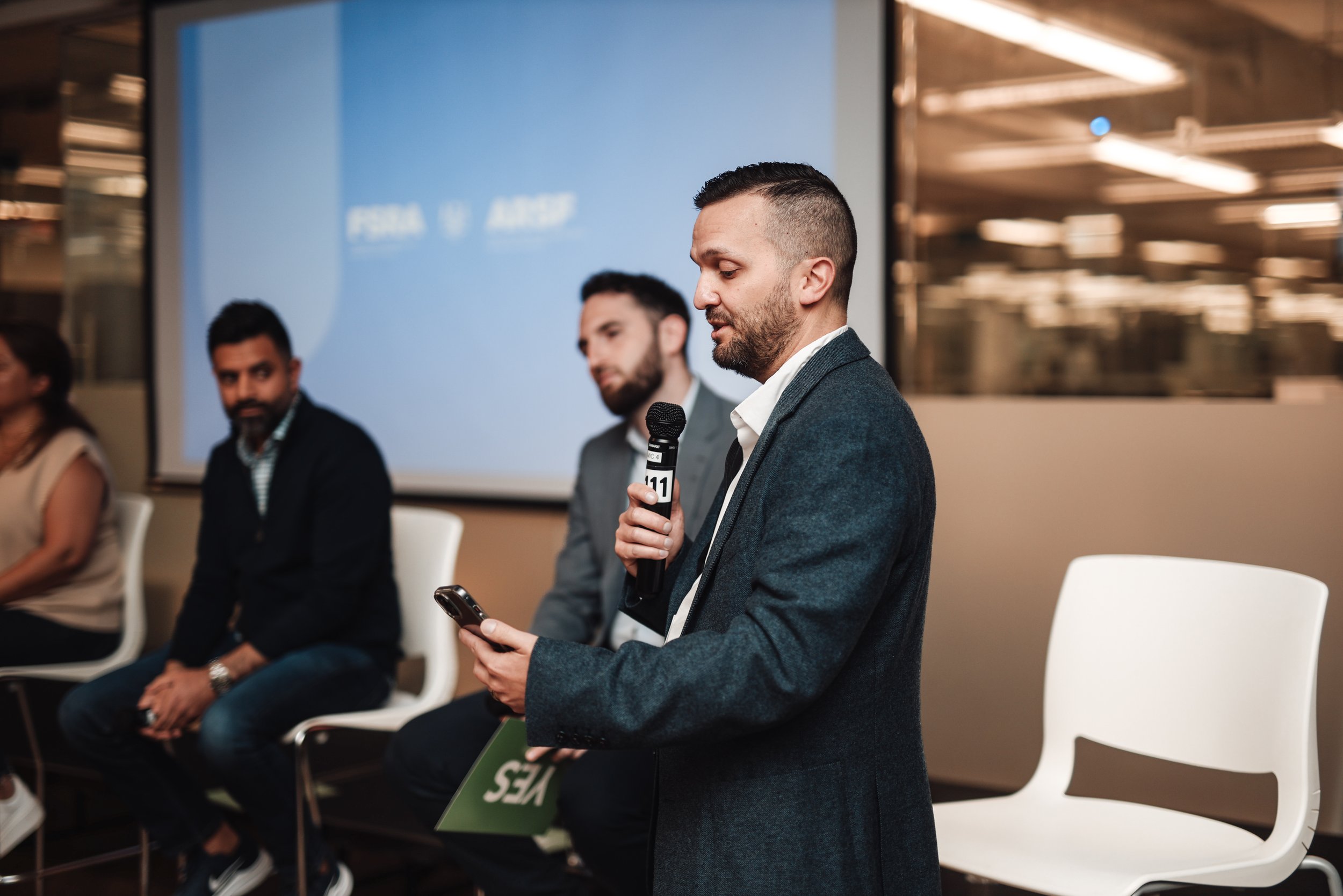 Man in a dark suit speaking into a microphone while looking at his phone, seated in front of a blue screen at a panel discussion event.