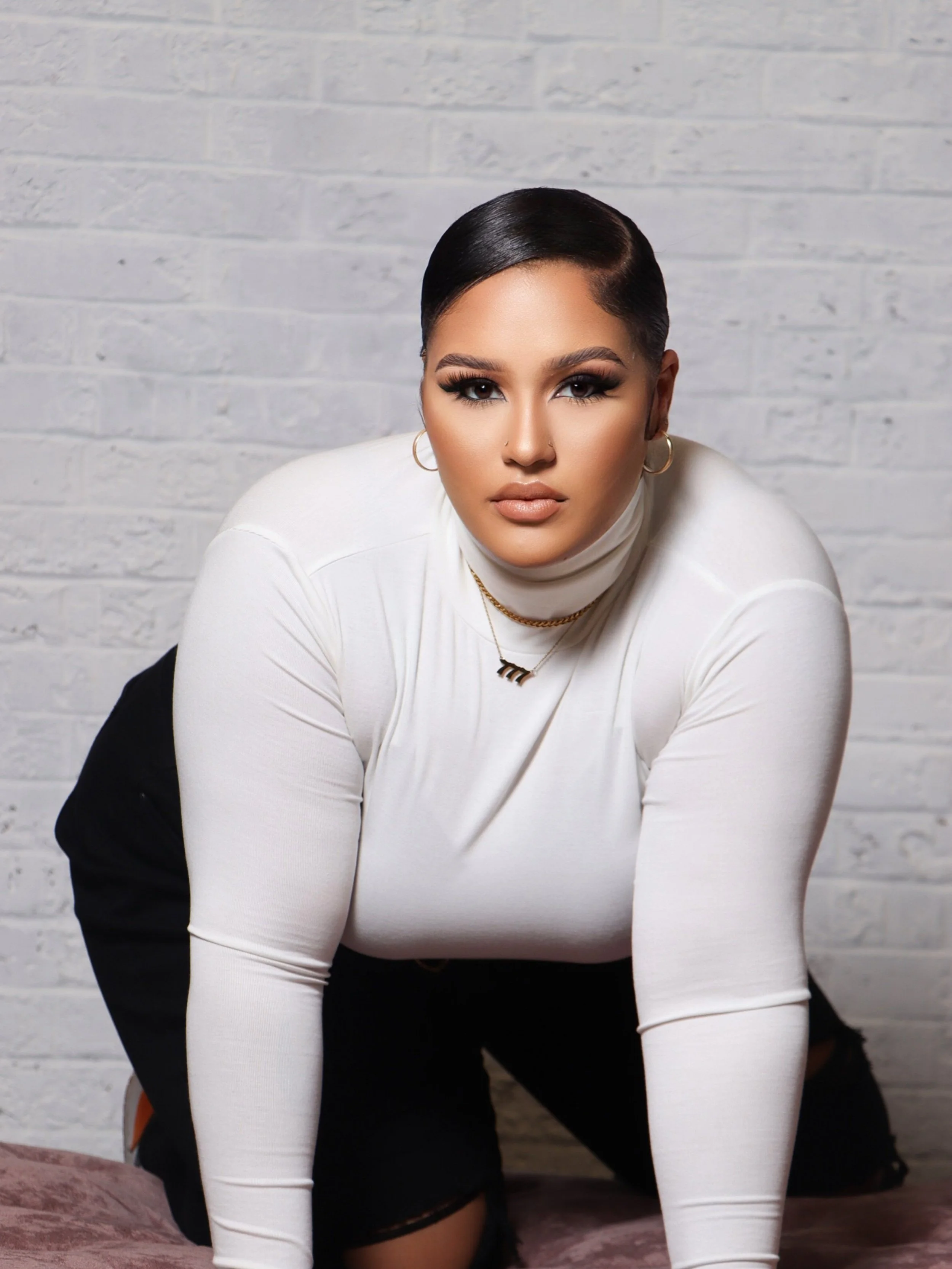 A woman with dark hair styled close to her head, wearing a white turtleneck top and black pants, poses against a white brick wall.