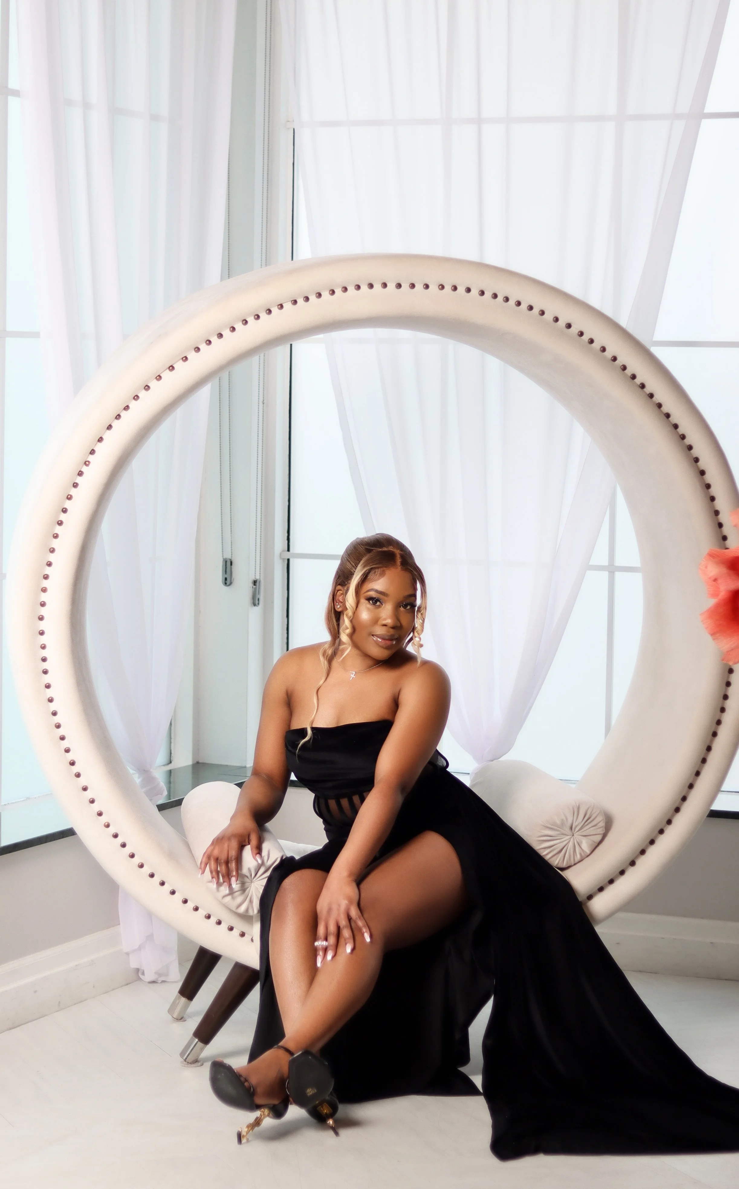 A woman posing in a black dress inside a minimalist, bright room with large windows and white curtains, seated on a modern, circular white chair with a cream cushion.