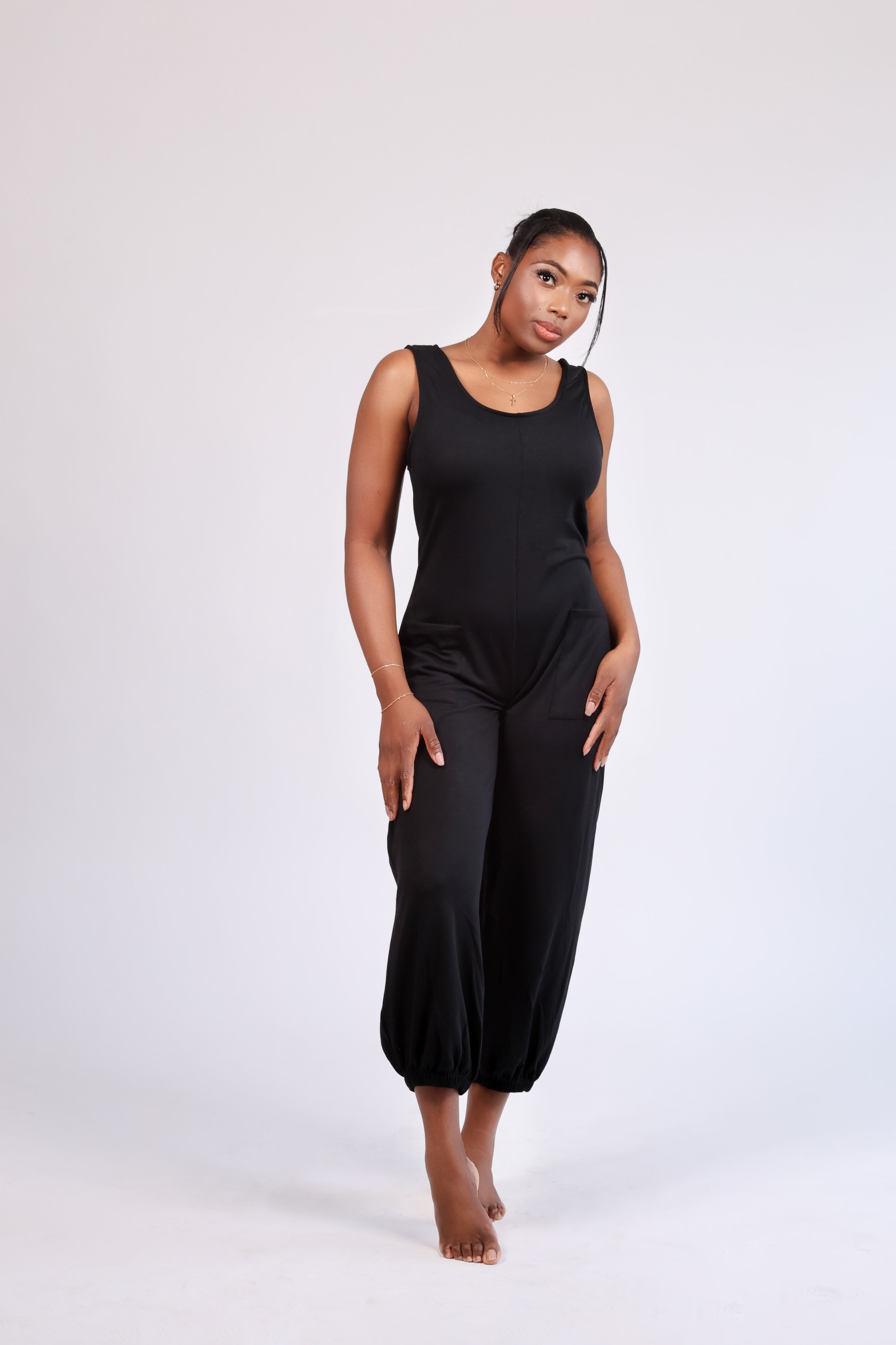 A woman in a black sleeveless jumpsuit standing barefoot against a plain white background.