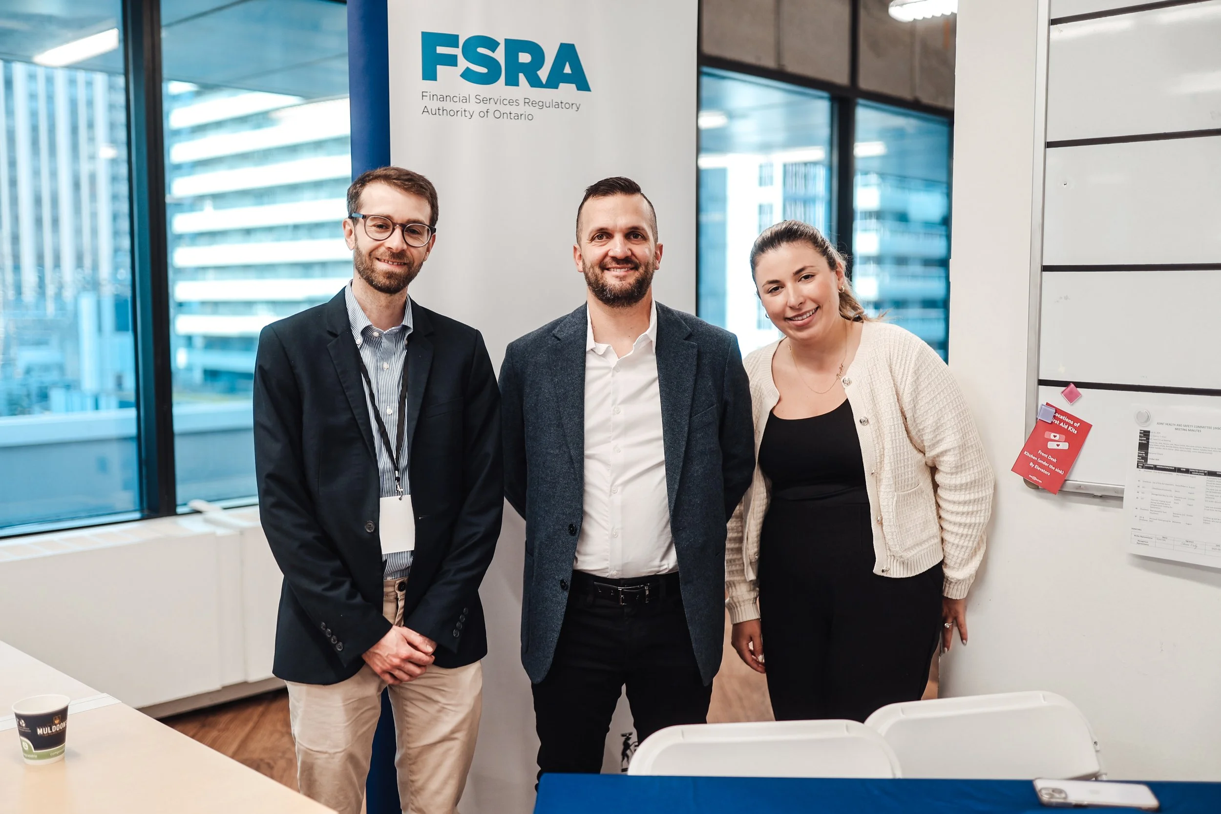 Three people standing indoors in front of a FSRA banner, smiling at the camera, with office windows showing city buildings in the background.