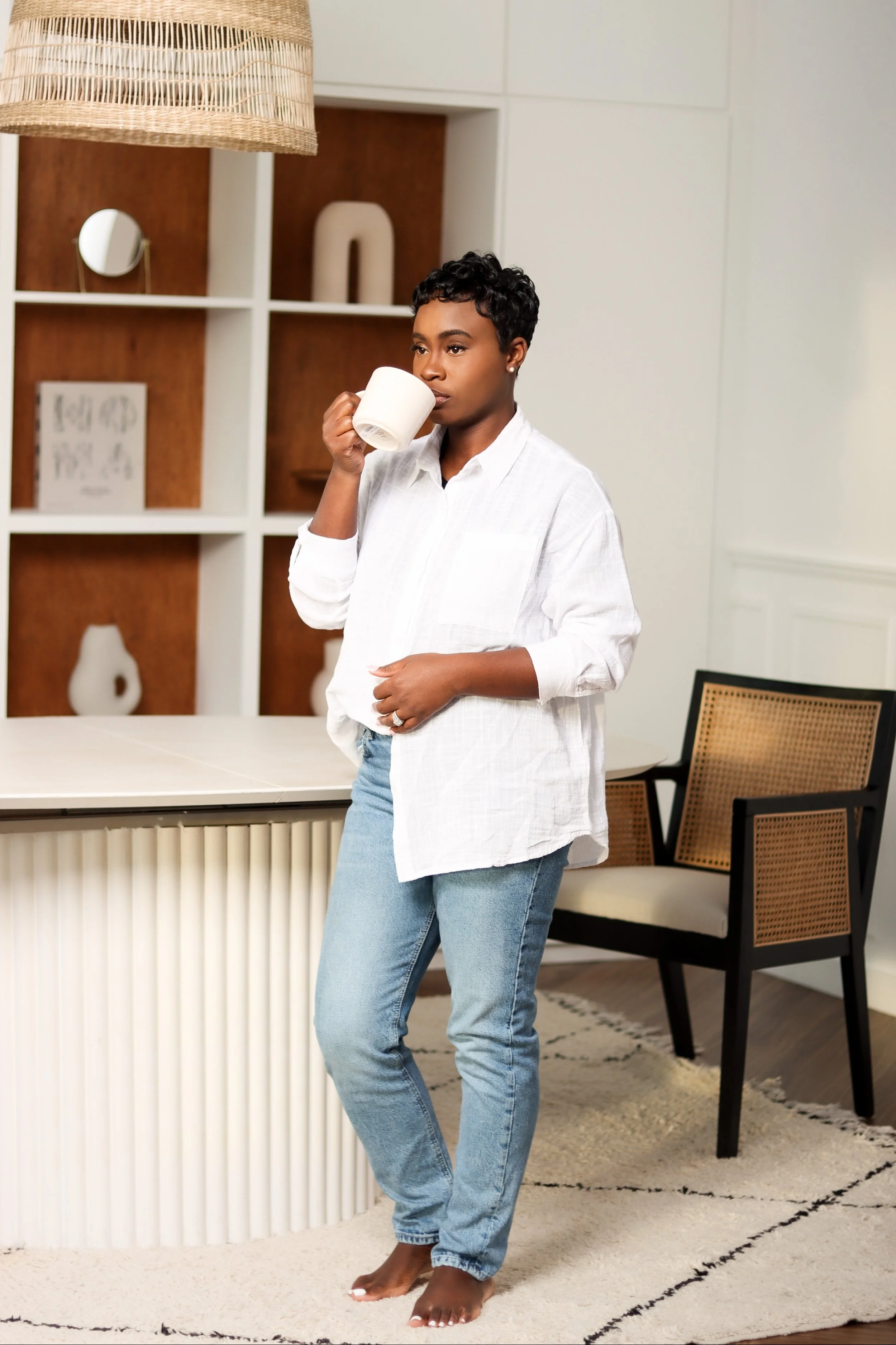 A woman with short curly hair drinking from a white mug in a modern living room with bookshelf and chair.