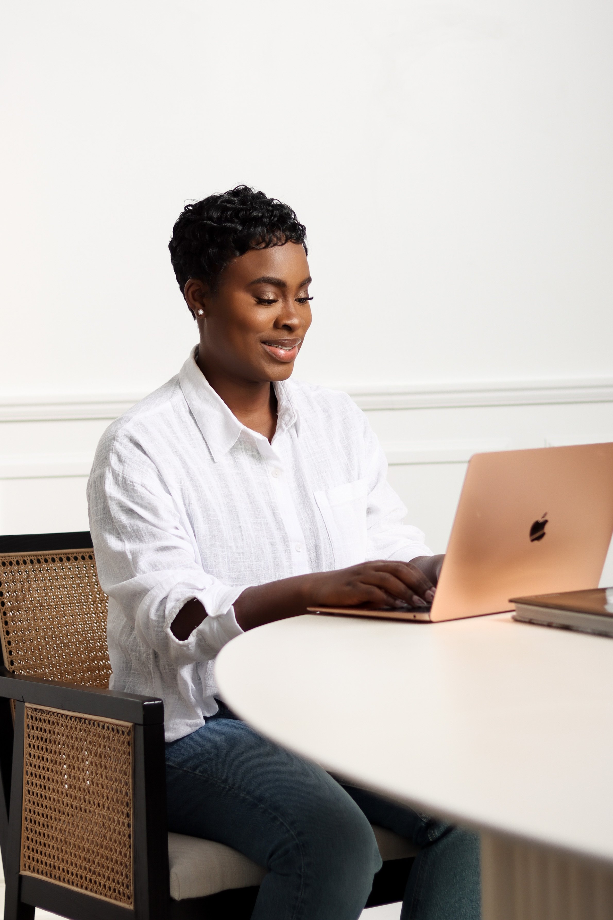 A woman sitting at a white table, working on a rose gold Apple MacBook laptop in a minimalist room.