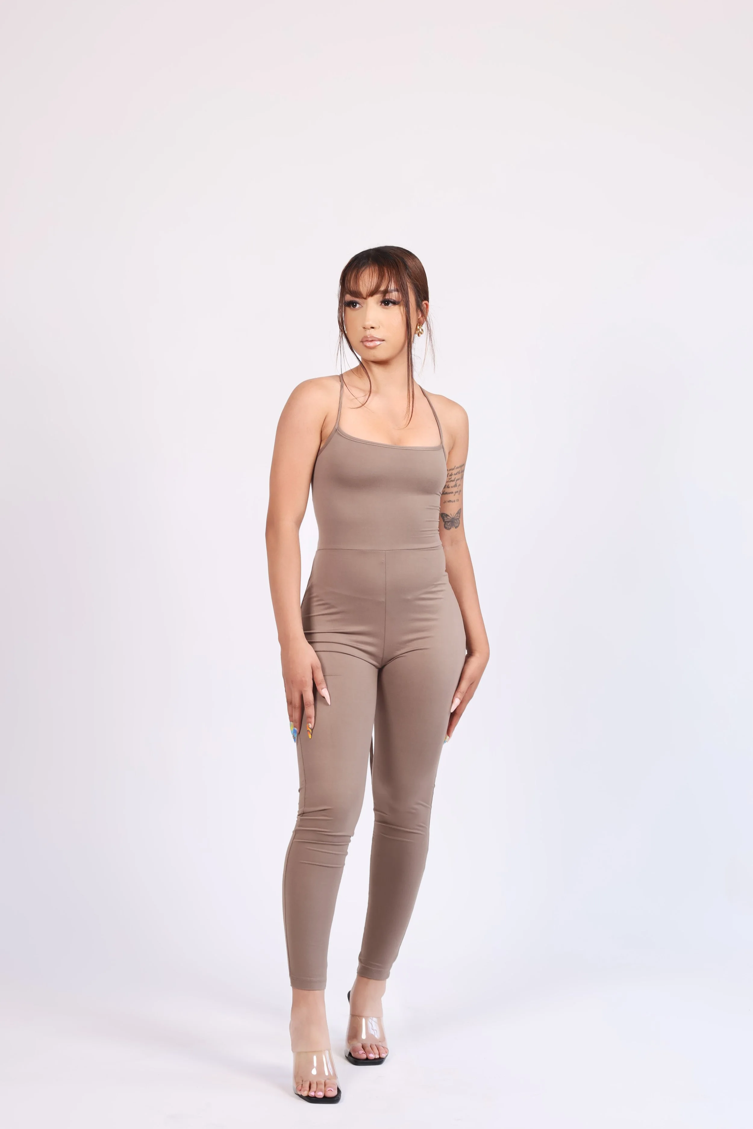 A woman wearing a matching taupe-colored tank top and pants, transparent high heels, standing against a plain white background.