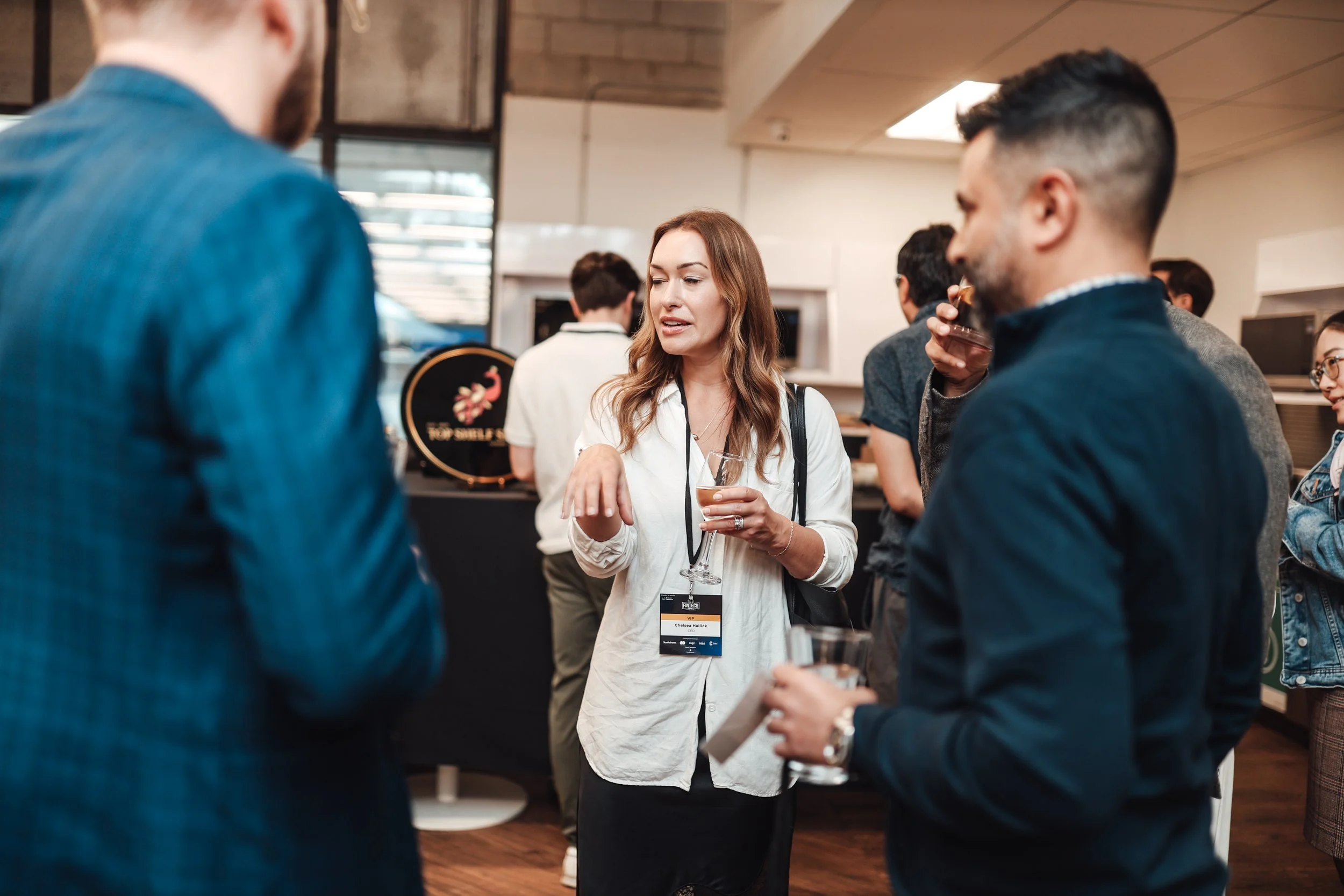Group of people socializing at an event or conference, with a woman in the center holding a drink and speaking to others.