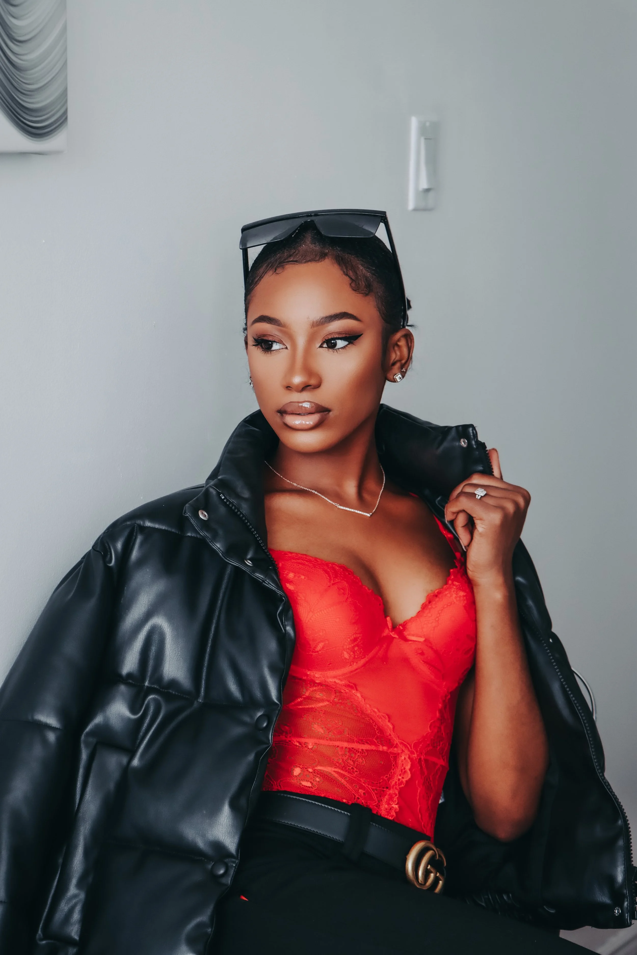 A stylish woman with short hair, wearing a black leather jacket over a red lace top, paired with black pants and a Gucci belt, posing against a plain wall with a light switch behind her.