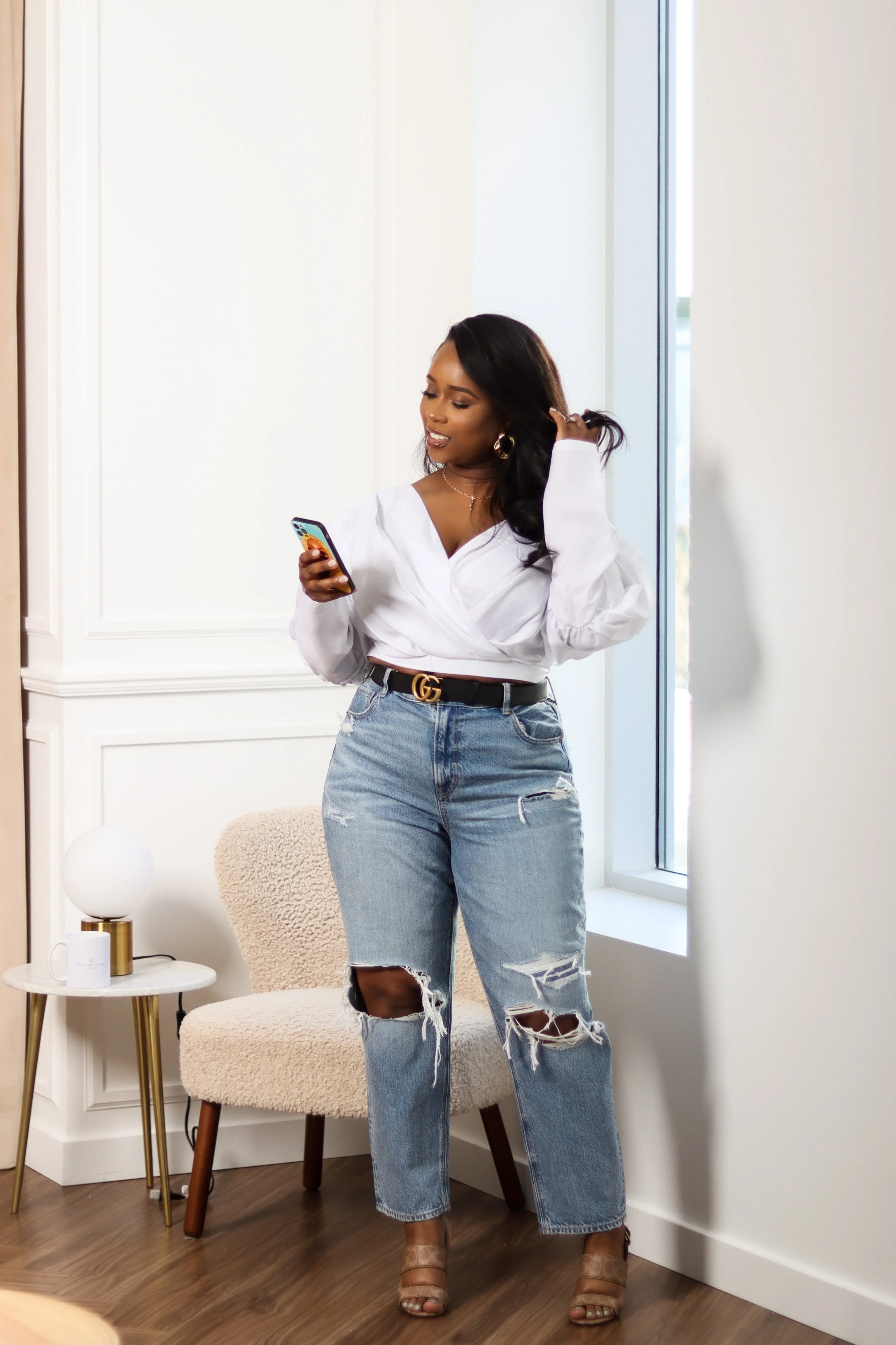 A woman with long dark hair, wearing a white blouse and ripped jeans, stands indoors near a window, looking at her phone.