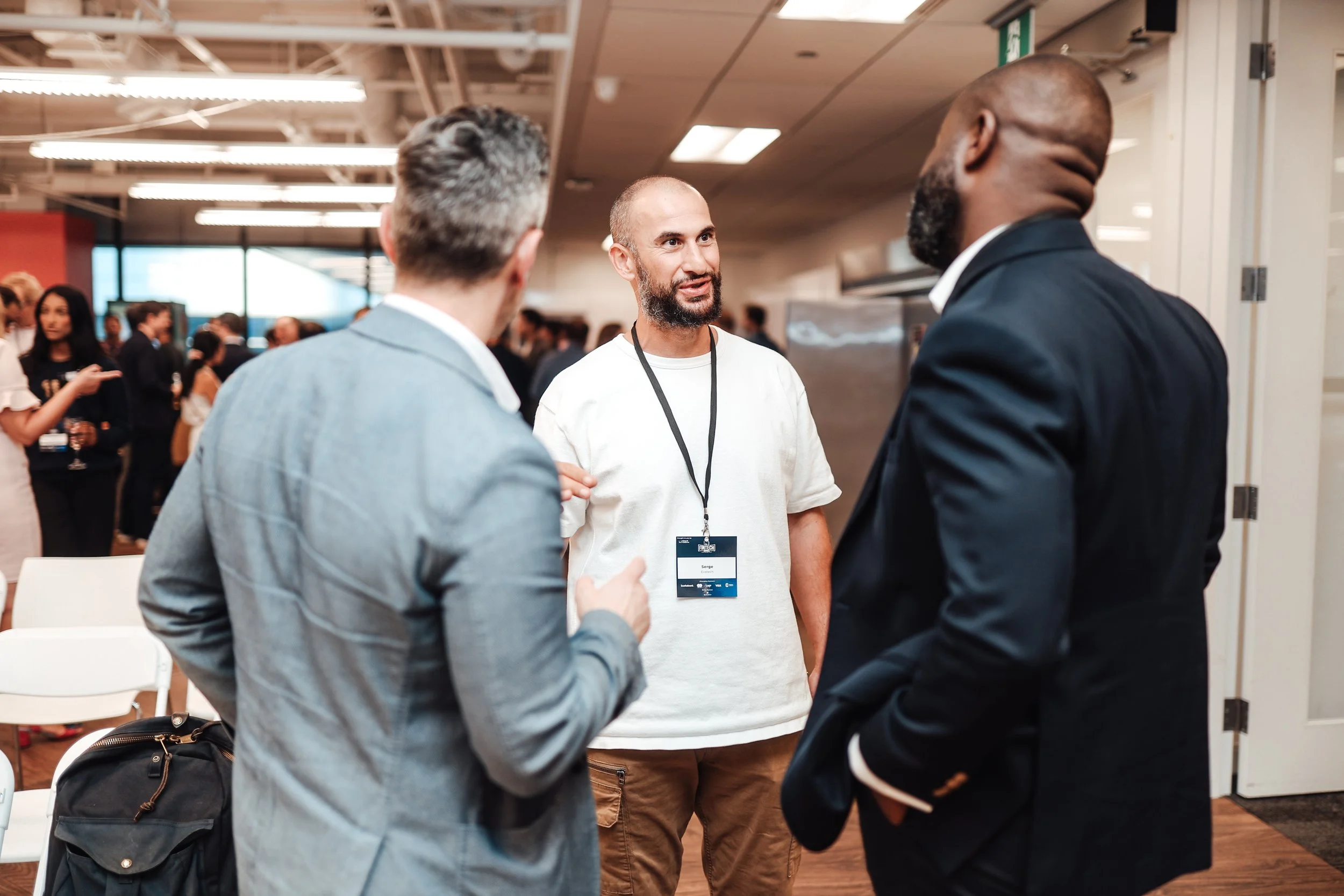 Three men having a conversation at a conference or networking event, with other attendees in the background.
